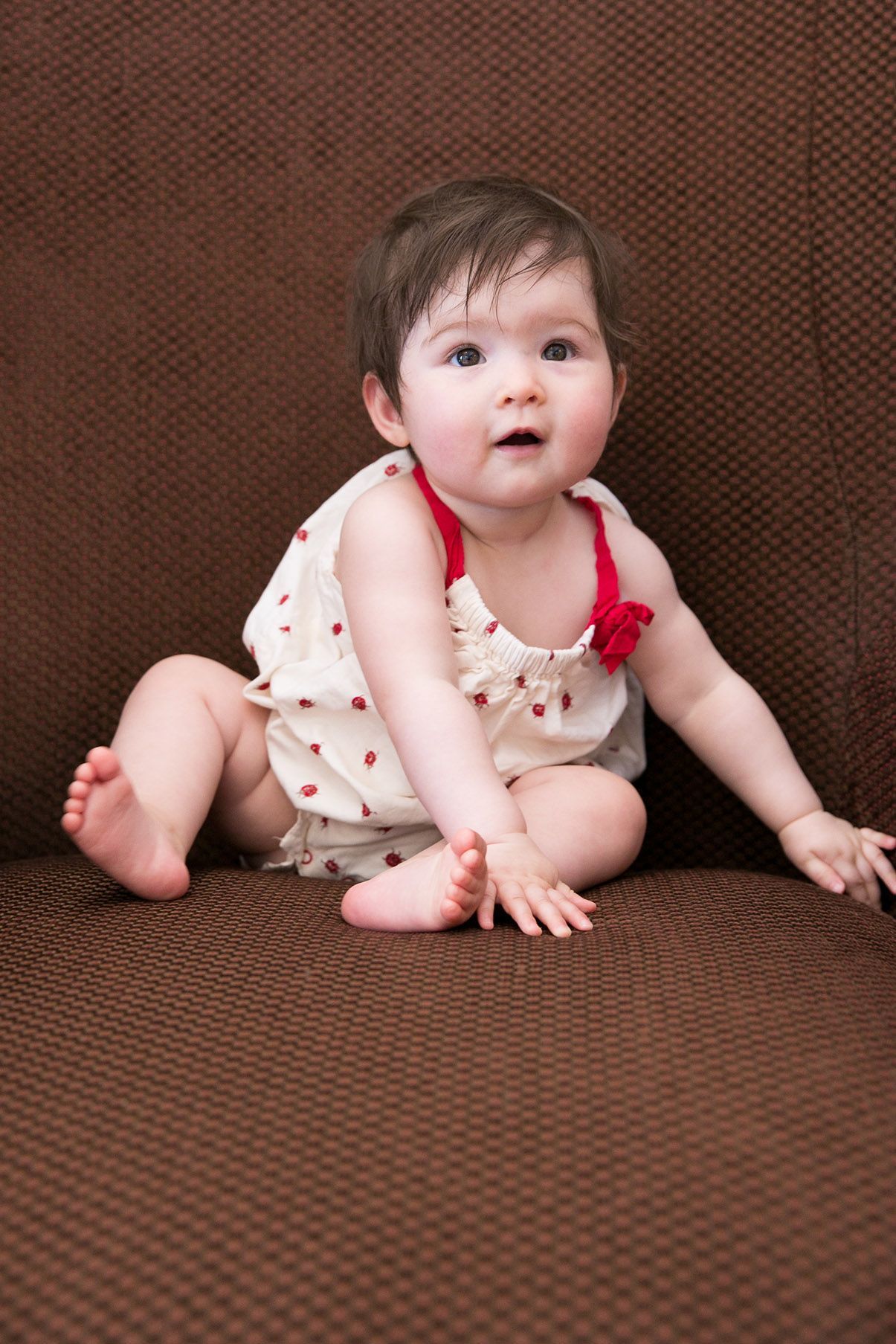 Baby in polka dot dress crawls on brown textured couch.