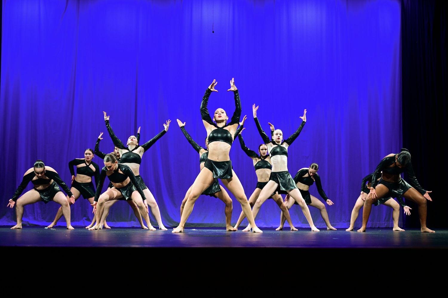 Dance performers in black costumes strike dynamic poses during a stage performance against a vibrant blue backdrop.