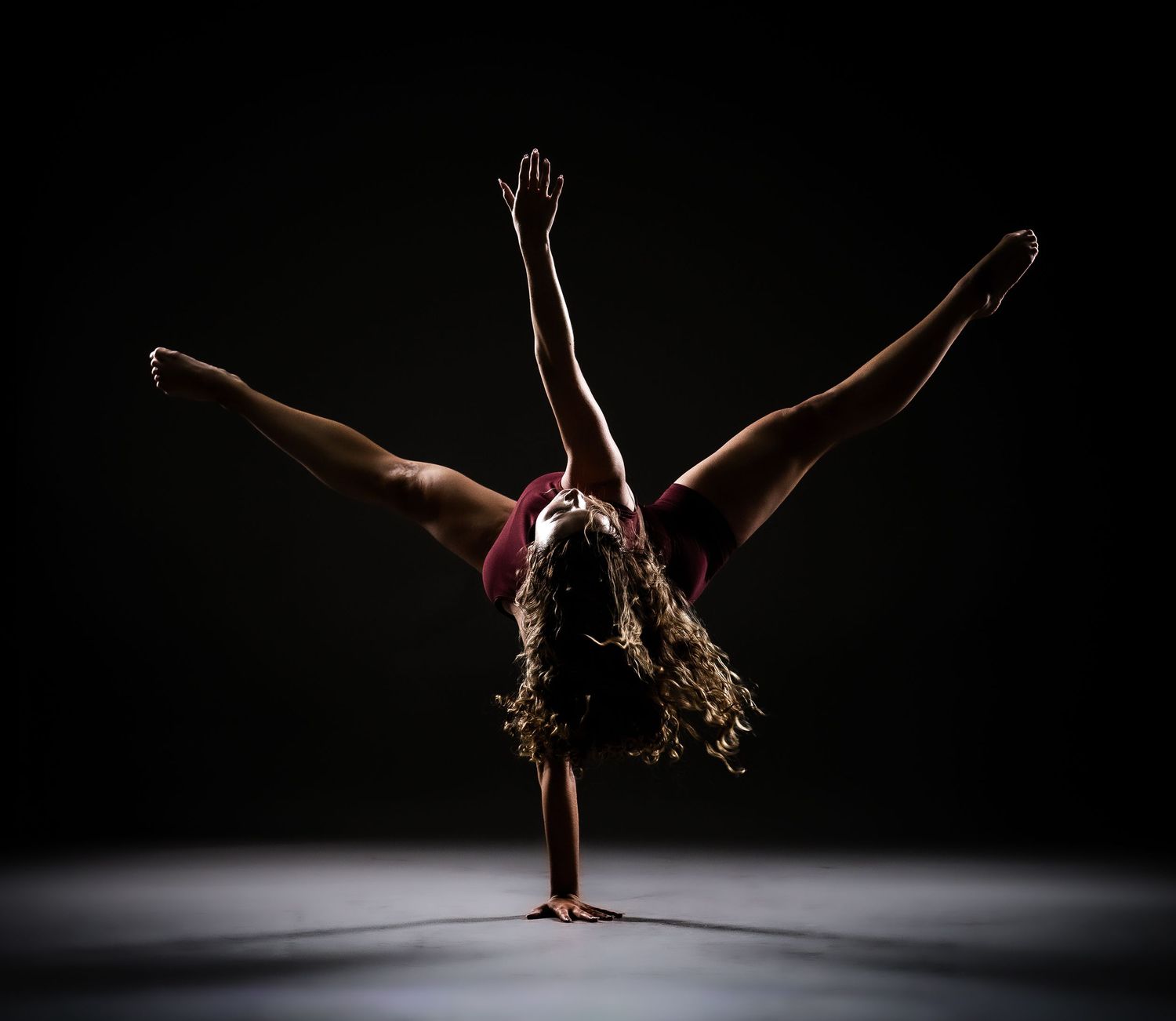 Gymnast performing a one-handed handstand split under dramatic spotlight on dark stage.