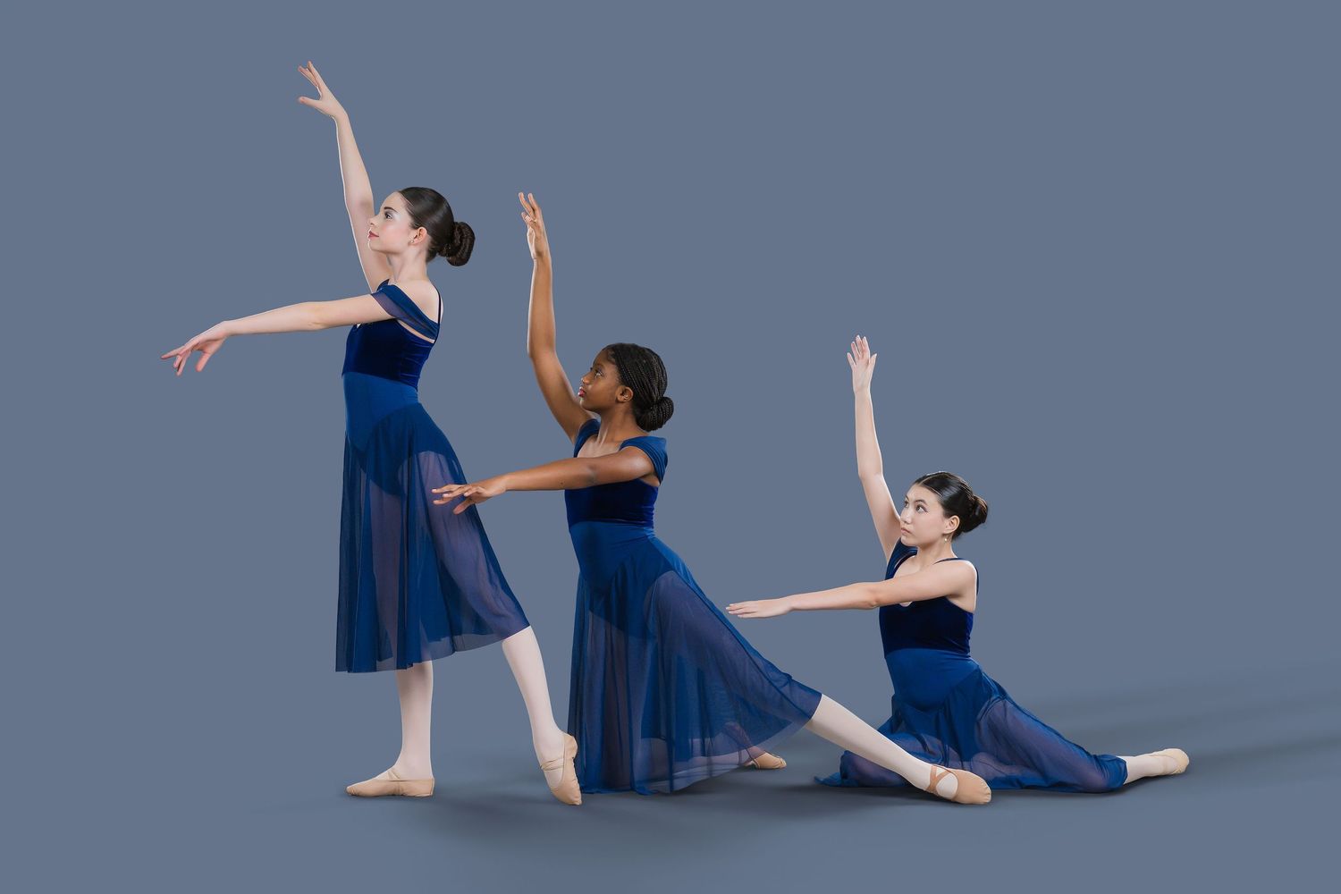 Three ballet dancers in blue leotards and skirts pose gracefully against a gray studio background.