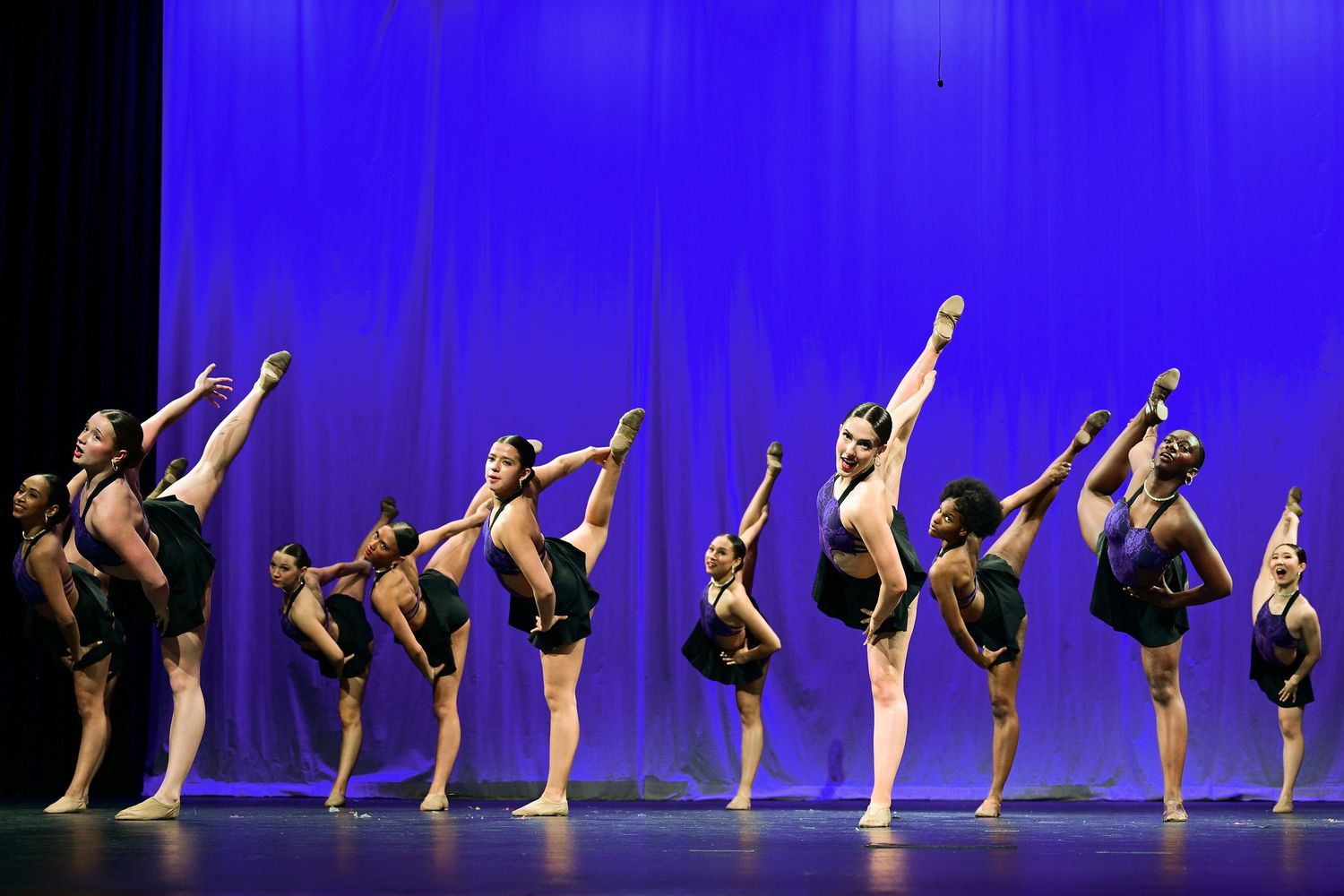 A line of dancers in black costumes perform synchronized high kicks on a blue-lit stage.