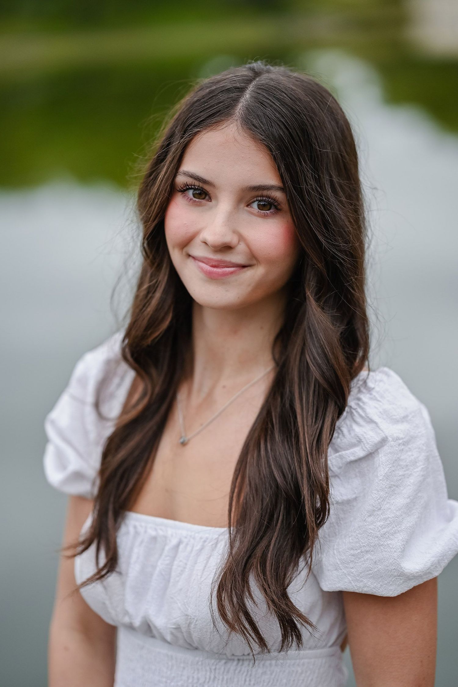 Woman in white peasant top poses outdoors by water in portrait series.