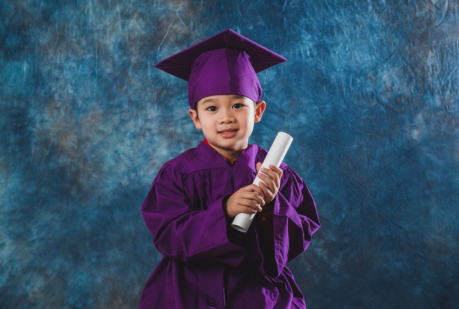 Child in purple graduation gown holds diploma while posing against blue background.