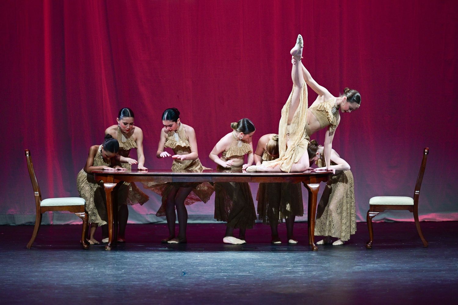 Performers in elegant costumes act out a dramatic scene on a dimly lit stage with a deep red backdrop.