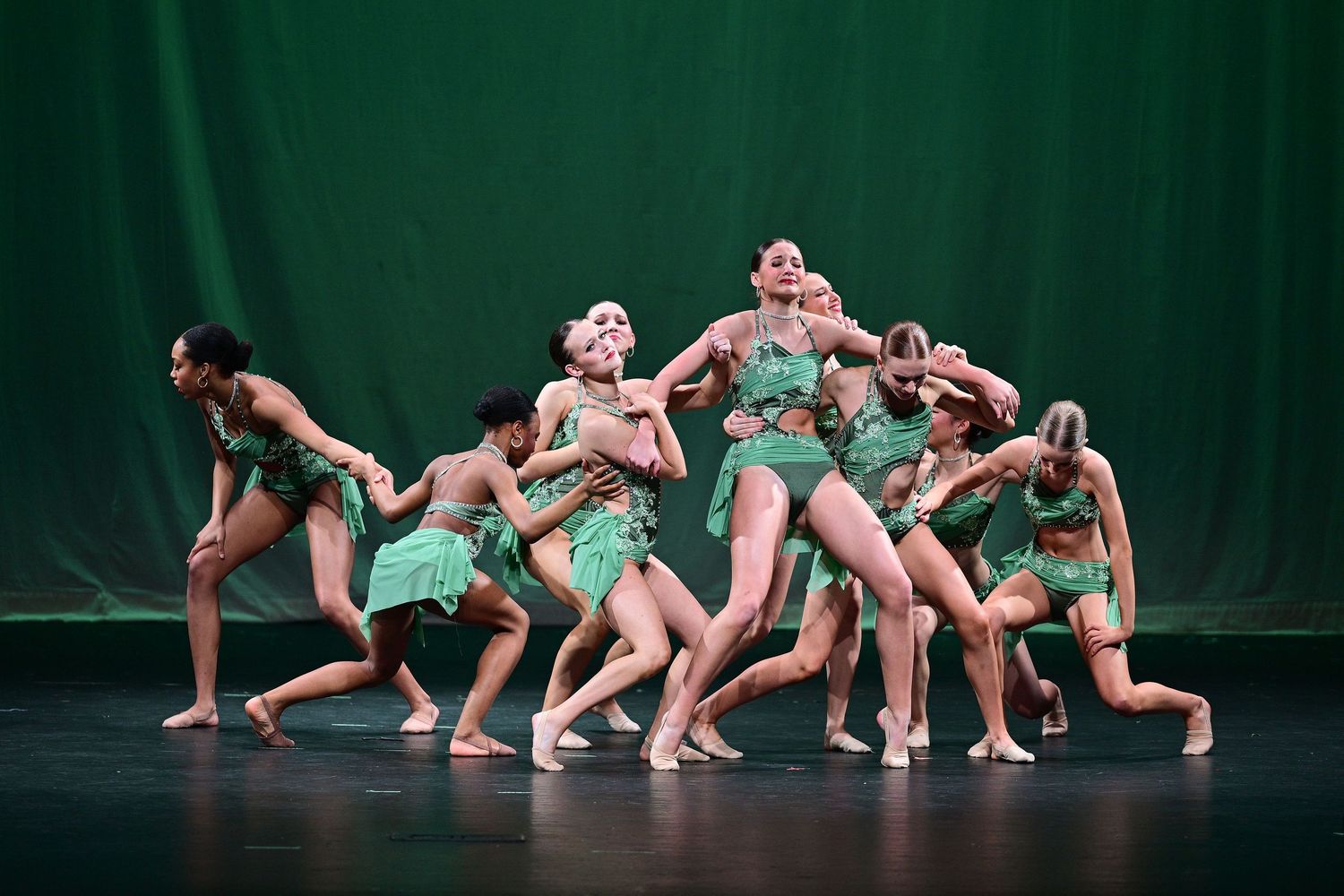 Dancers in green costumes perform a dynamic group routine on a stage with a green backdrop.