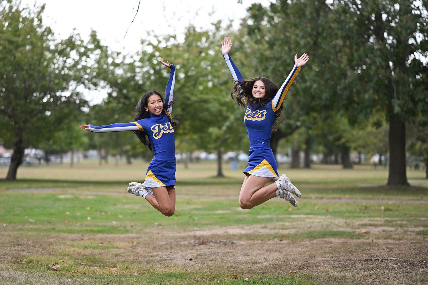Cheerleaders in blue uniforms perform synchronized jumps in a grassy outdoor field.