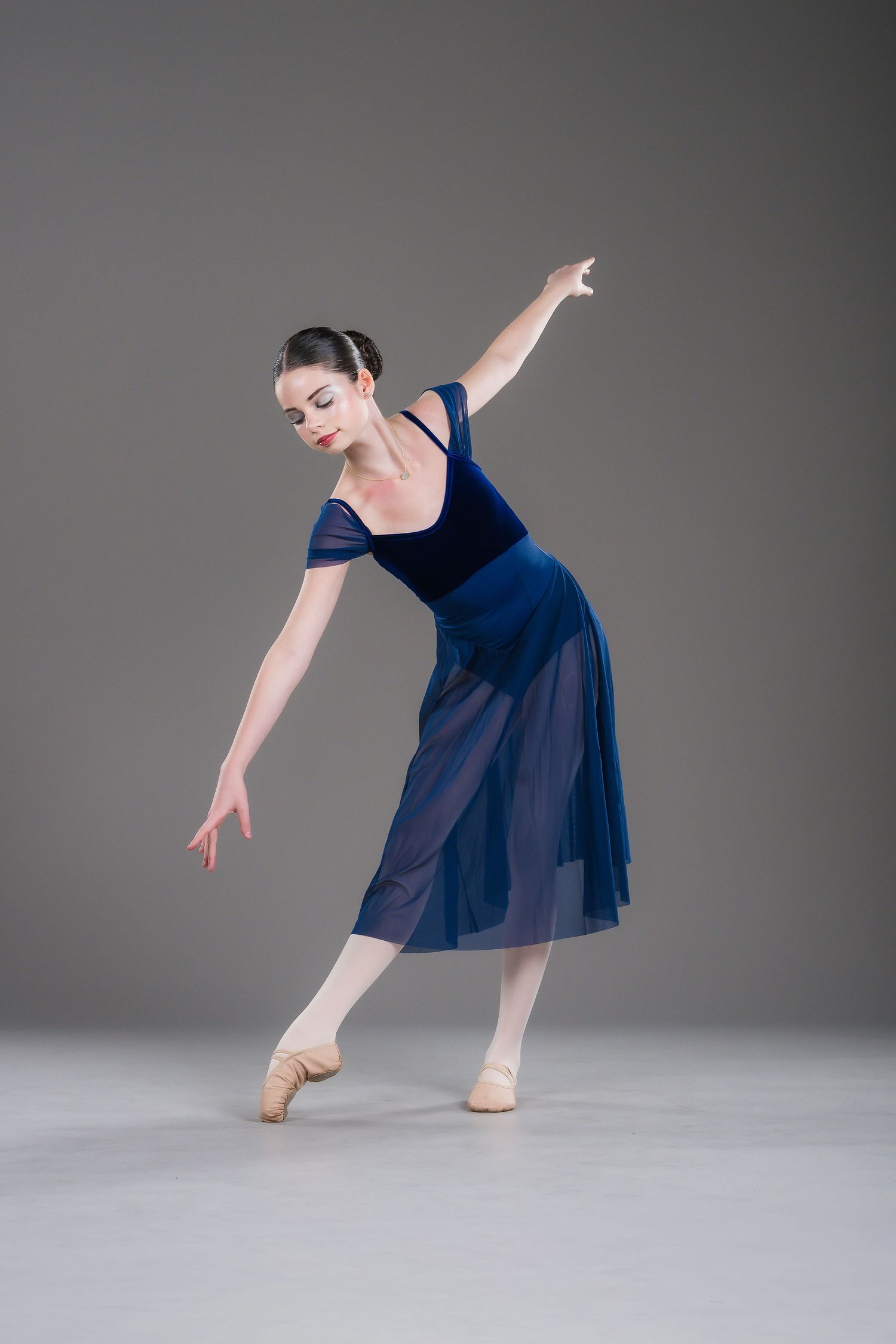 Elegant ballerina in blue velvet dress performing graceful arabesque pose on gray studio background.