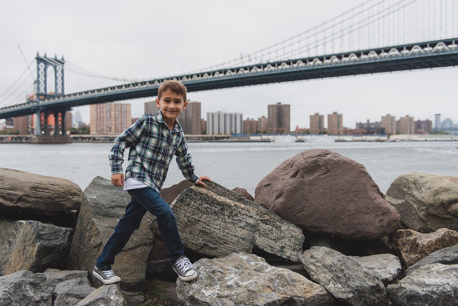 Couple sitting on large rocks by Manhattan Bridge with NYC skyline in background.
