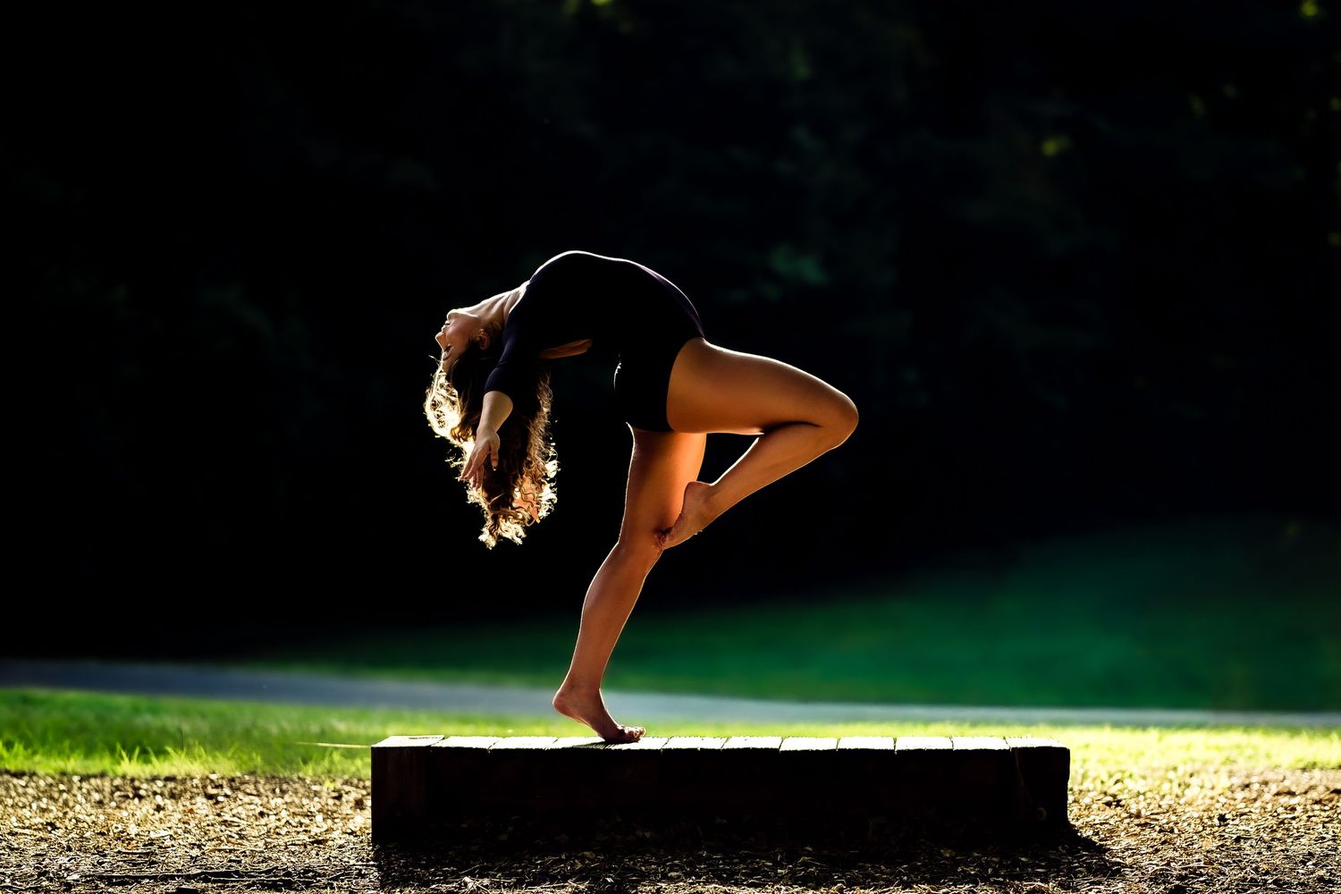 Woman performing a graceful yoga balance pose outdoors on a stone platform, silhouetted against lush green trees.