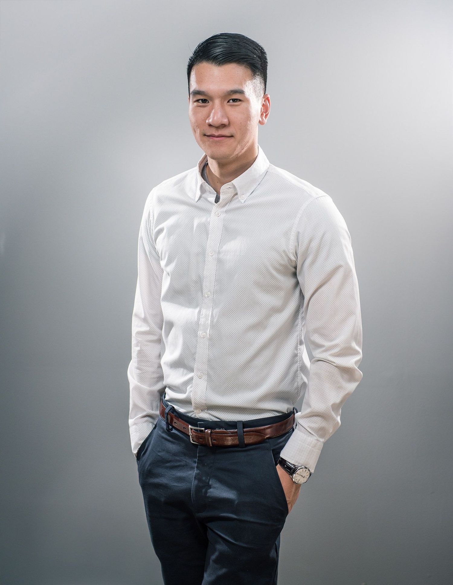 Professional portrait of man in white button-down shirt and dark pants against grey background.