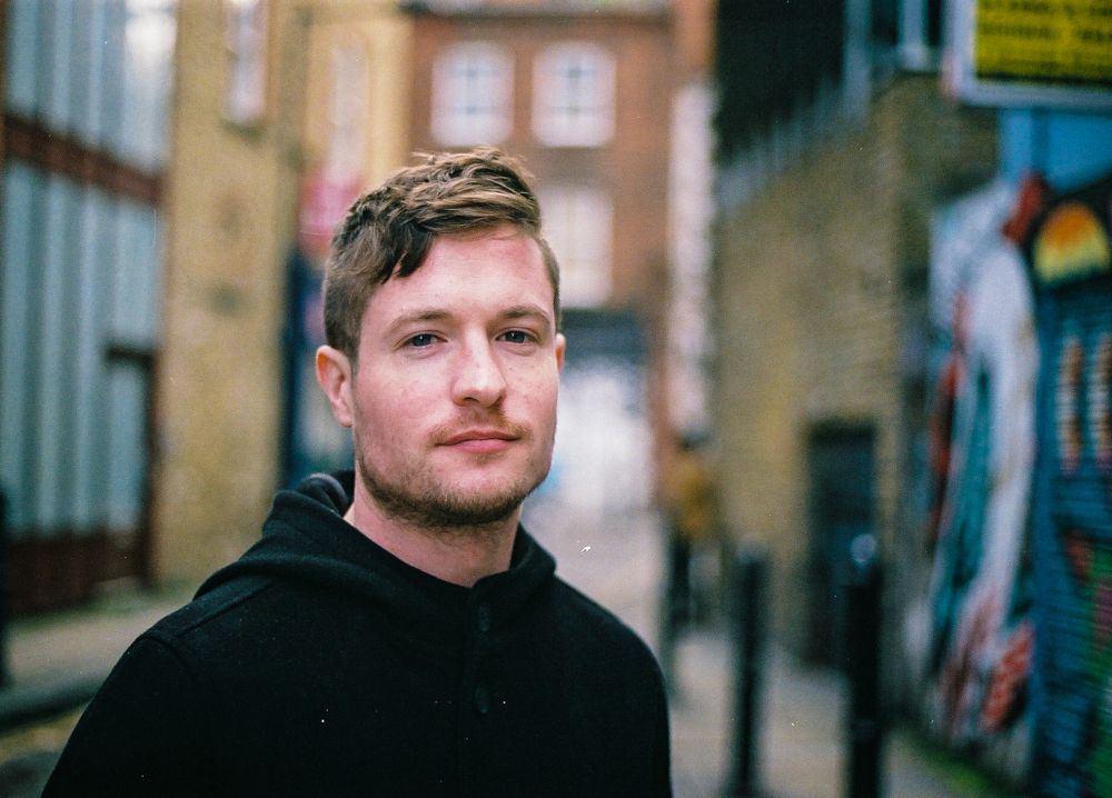 Corporate outdoor portrait of a man in his twenties wearing a dark coat against an urban background in Brick Lane, London