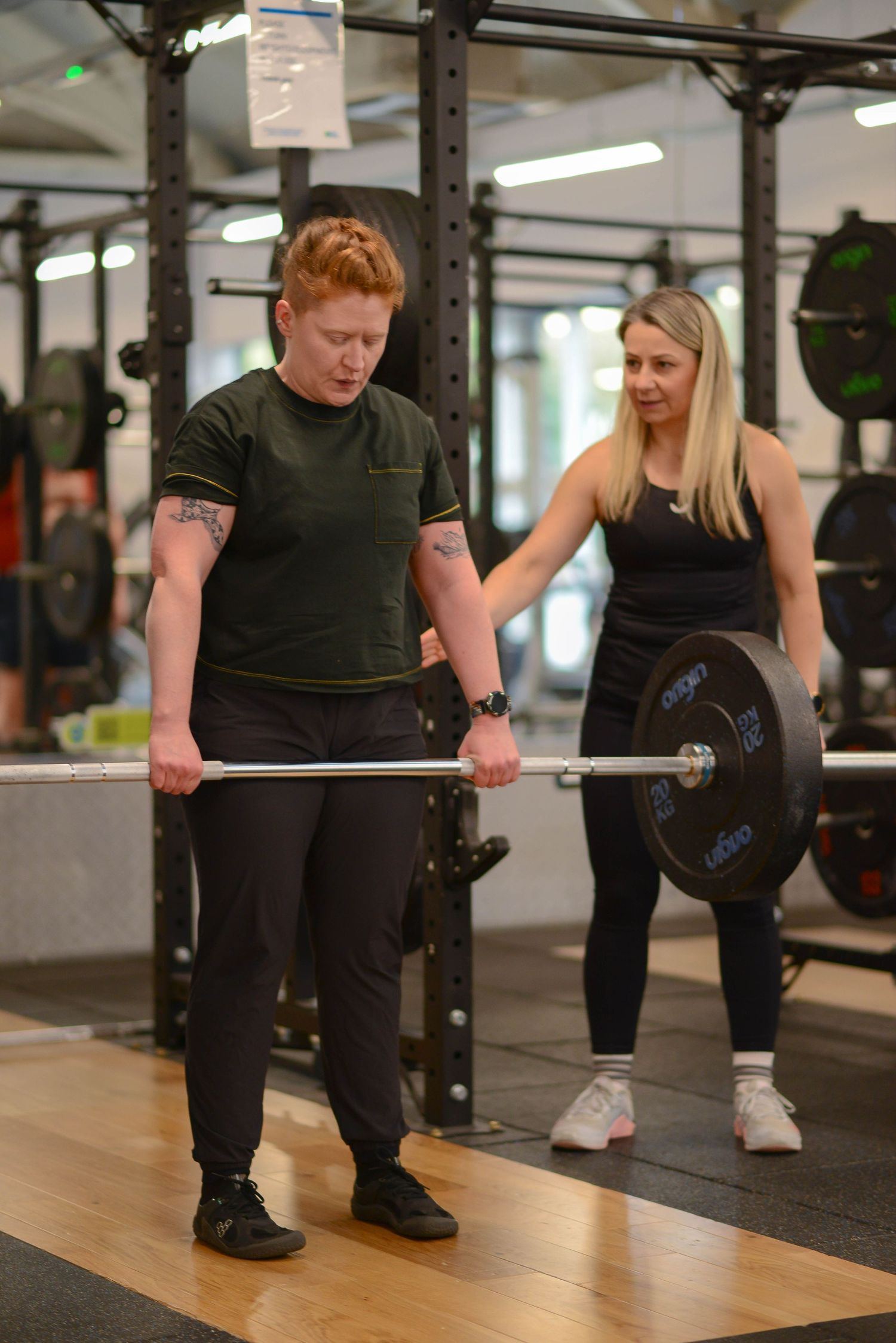 Glasgow portrait of two women in a gym, one with red hair and one with blonde hair, the latter training the former to deadlift a barbell