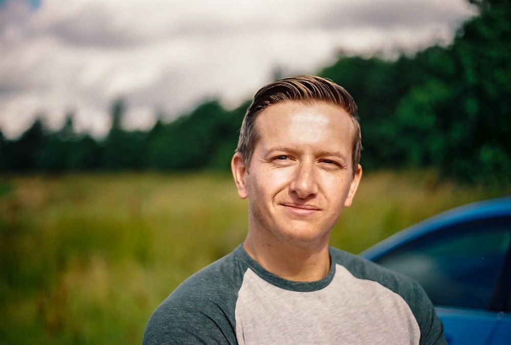 Corporate outdoor portrait of a smiling man in his thirties wearing a t-shirt against a background of an open field surrounded by trees