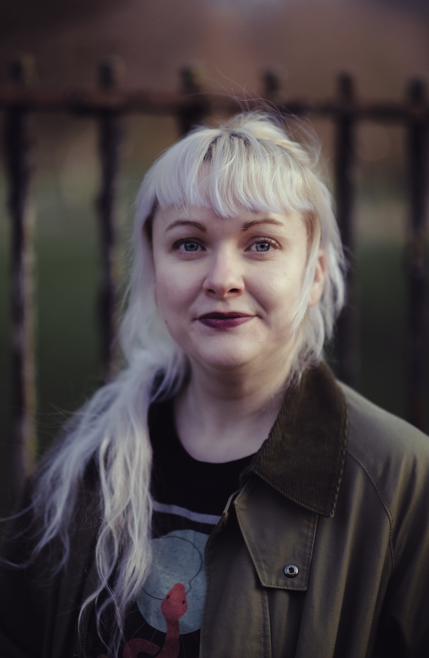 Glasgow creative headshot of a woman in her thirties with white hair and a green jacket in front of a black iron fence of a park