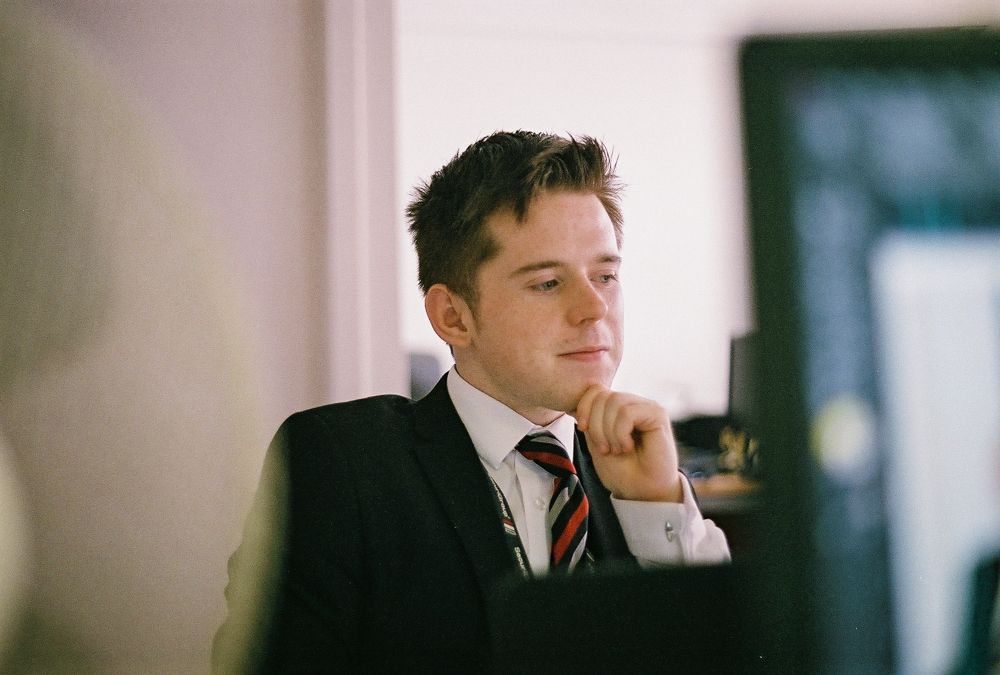 Corporate portrait of a smiling man in his twenties wearing a black suit, white shirt and striped tie in an office environment