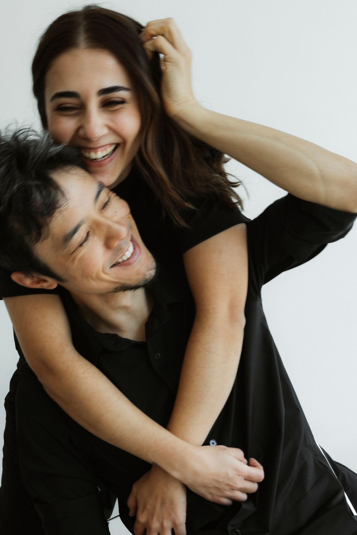 A joyful couple in black outfits laughing and embracing, with the woman hugging the man from behind against a white background.