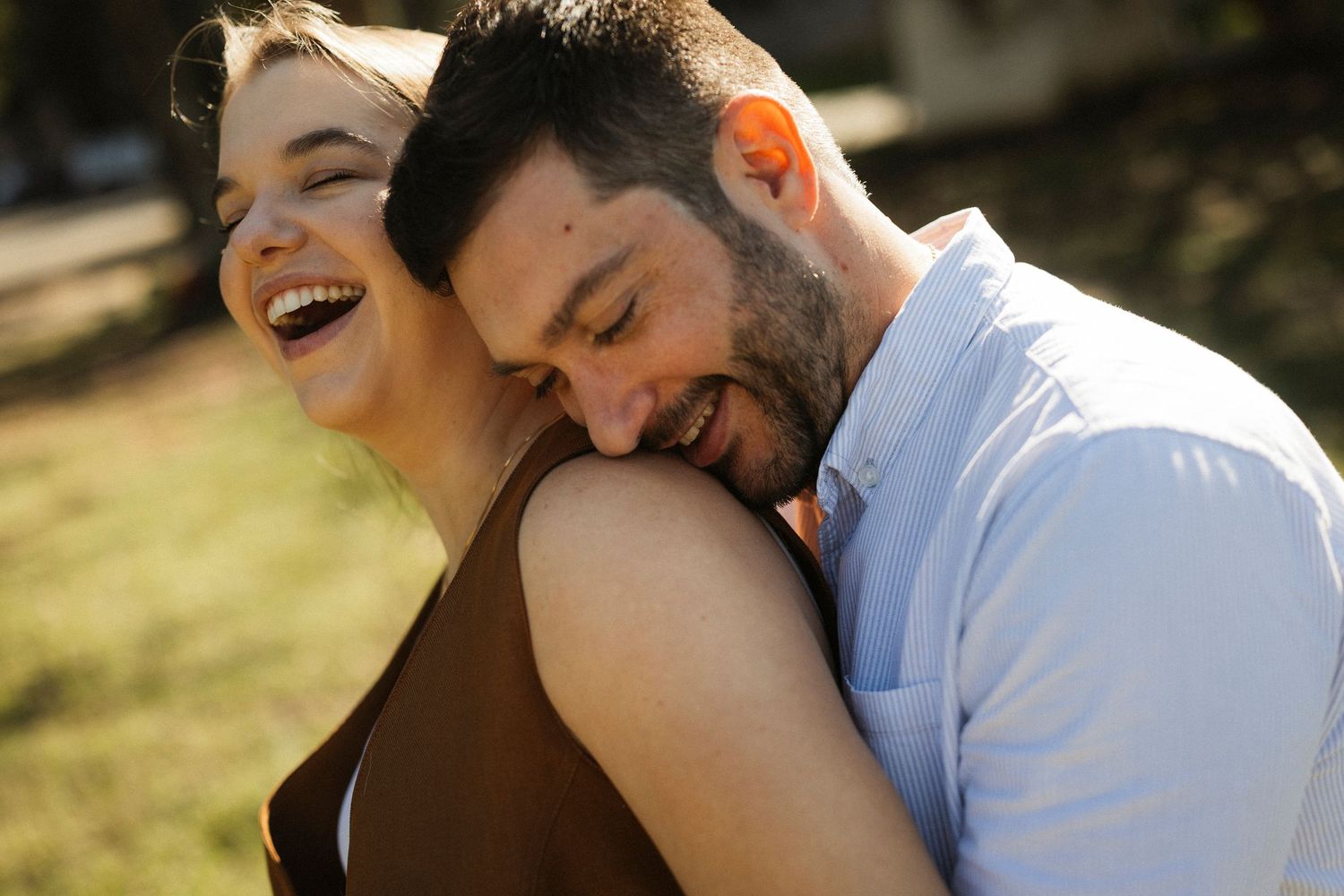 Happy couple embracing outdoors in golden sunlight, woman laughing joyfully as man smiles with eyes closed.