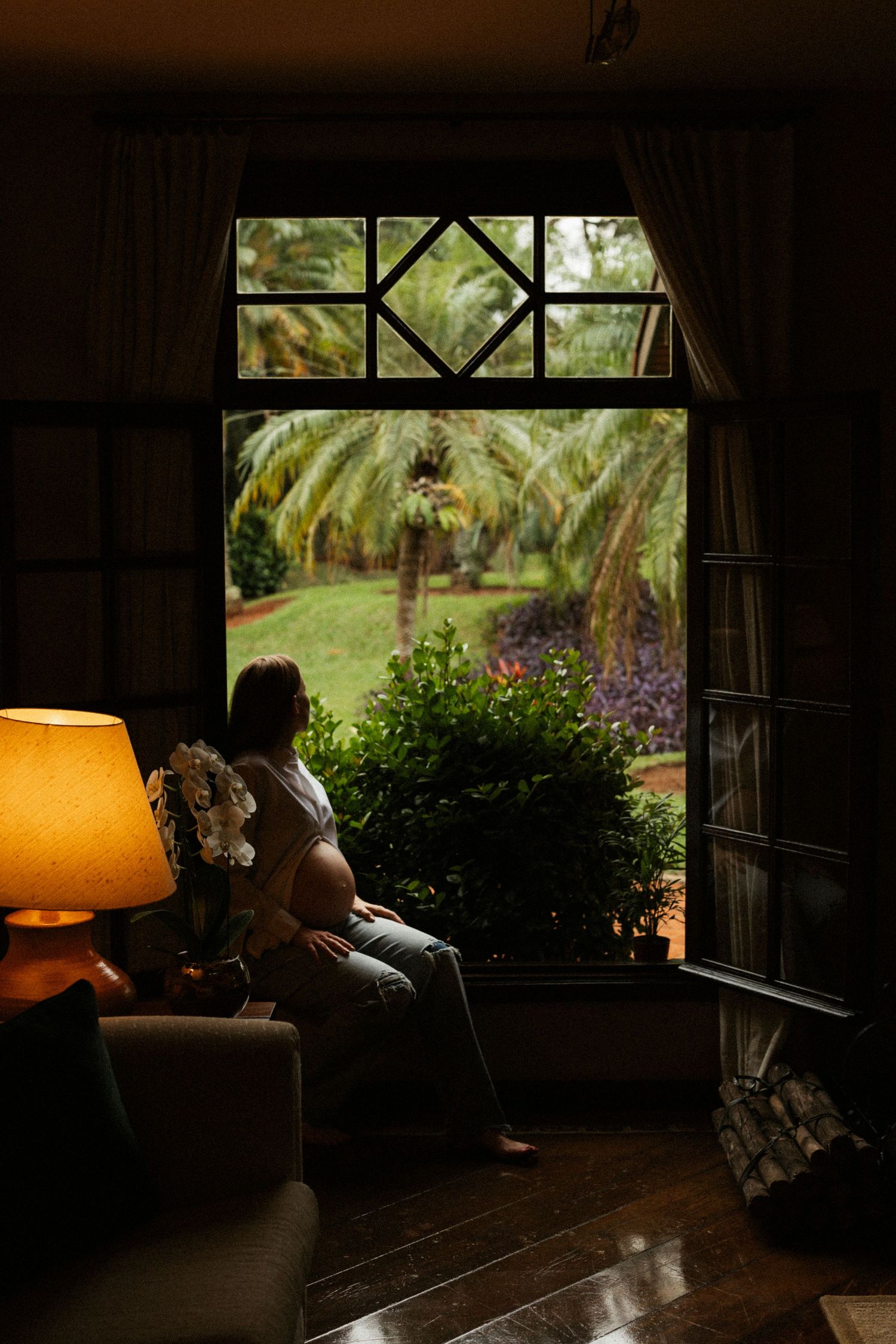 Person relaxing in cozy armchair by window overlooking lush garden with palm trees and purple flowers.