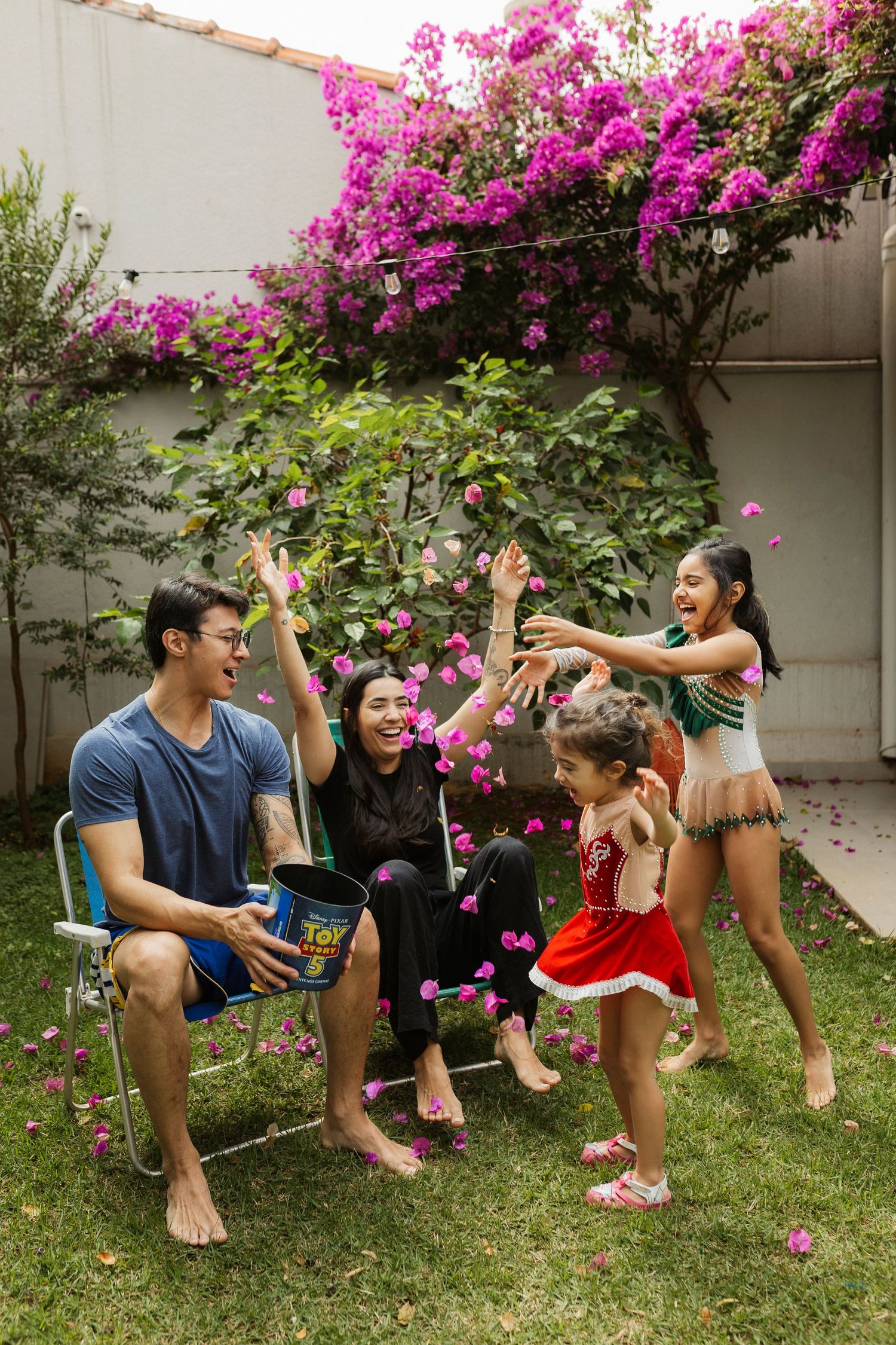Family playing outdoors in garden with pink bougainvillea flowers, tossing petals joyfully on green lawn.