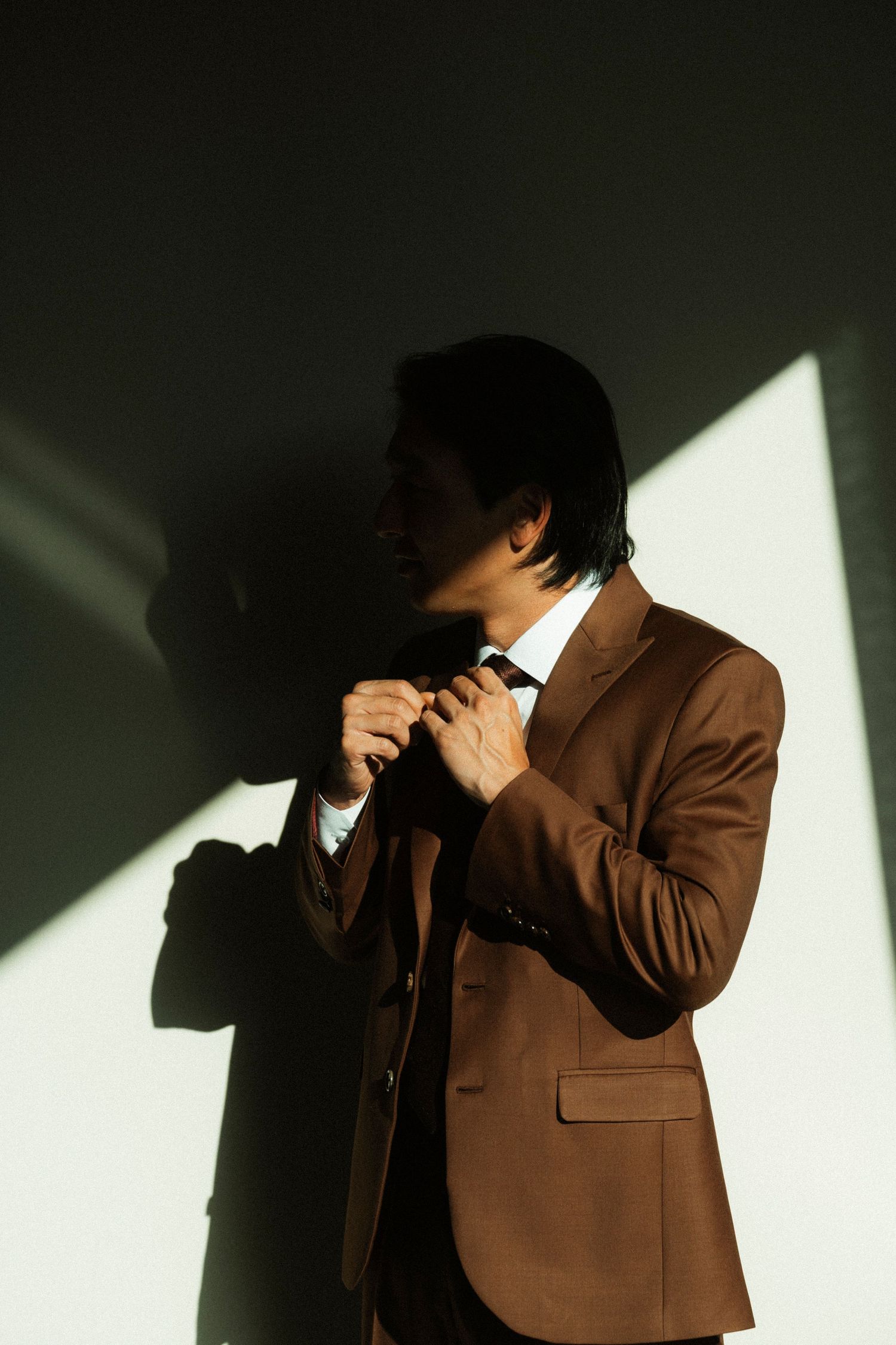 Man in brown suit adjusting tie in dramatic sunlight against white wall.