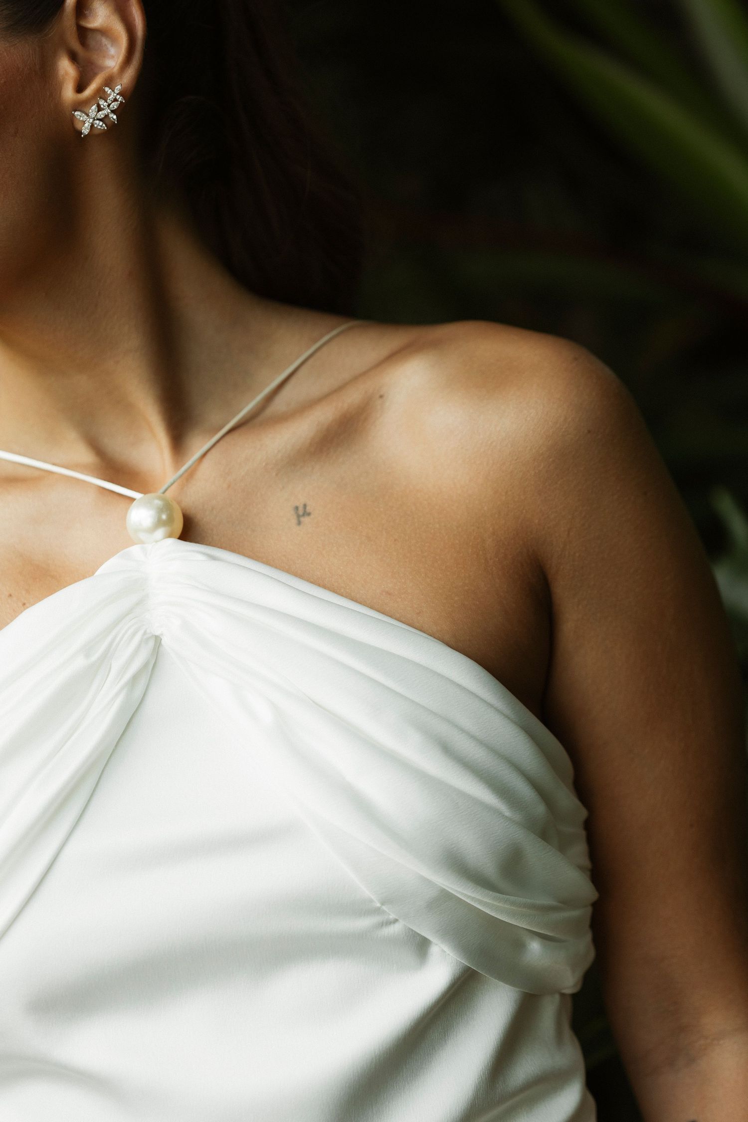 Woman in white halter dress with pearl detail, wearing diamond earrings and small collarbone tattoo.
