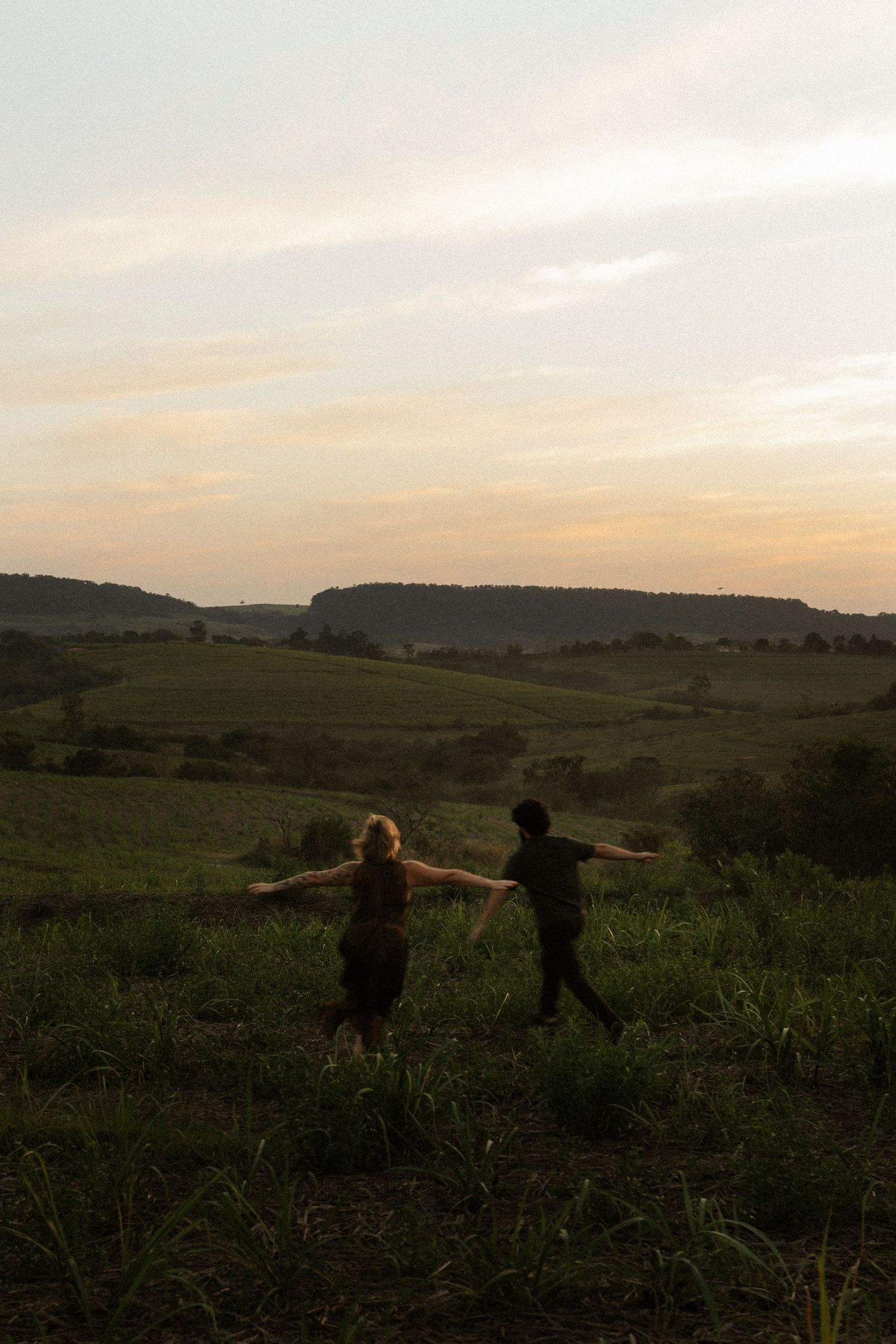 Two people stand with arms outstretched in a lush green field at sunset with rolling hills in the background.