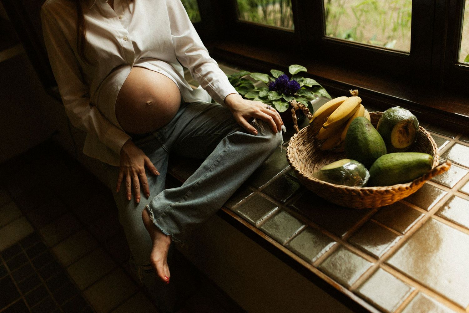Woman breastfeeding baby near window with basket of fresh fruits and vegetables on wooden table.
