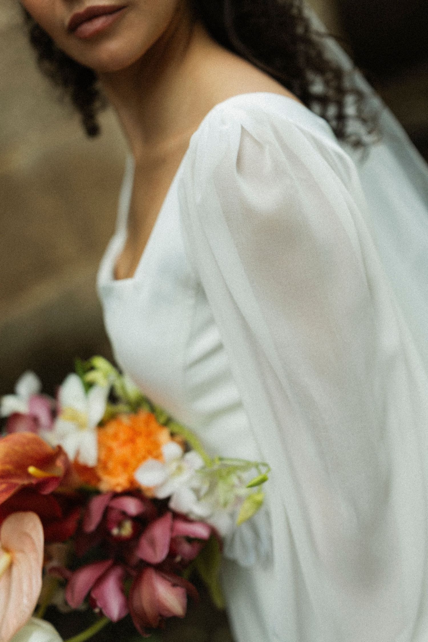 Bride in white long-sleeve gown holding colorful bouquet with orange, pink, and white flowers.