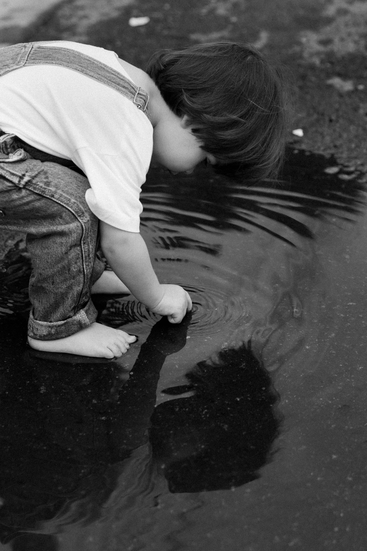 Black and white photos of someone leaning over to look at their reflection in a puddle.