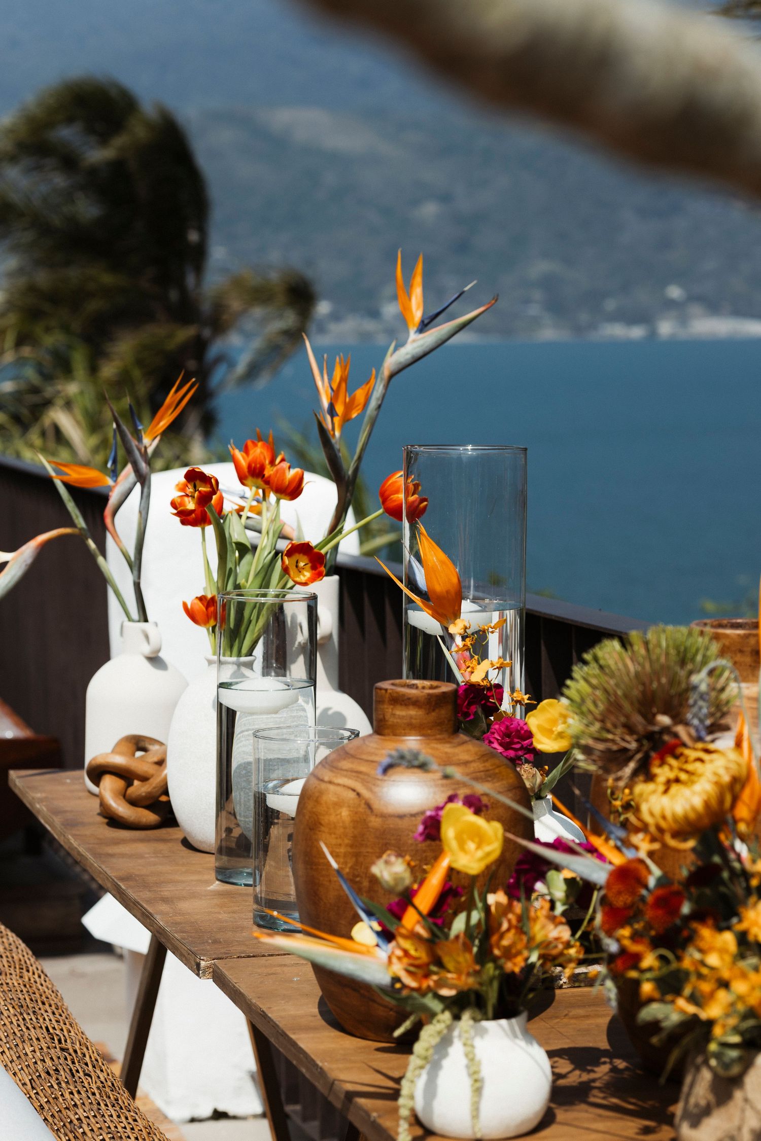 Tropical floral centerpieces with orange birds of paradise and white pumpkins on a wooden table overlooking the ocean.