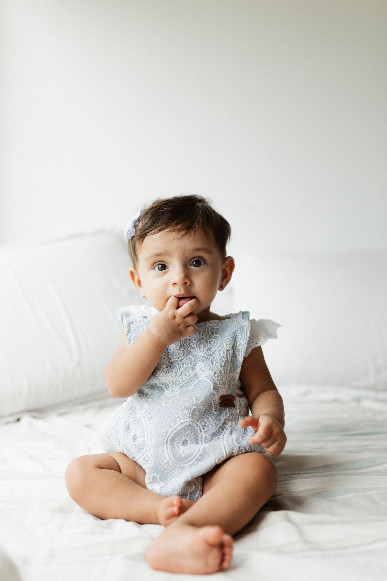 Baby in white lace dress sitting on white bedding against neutral background.
