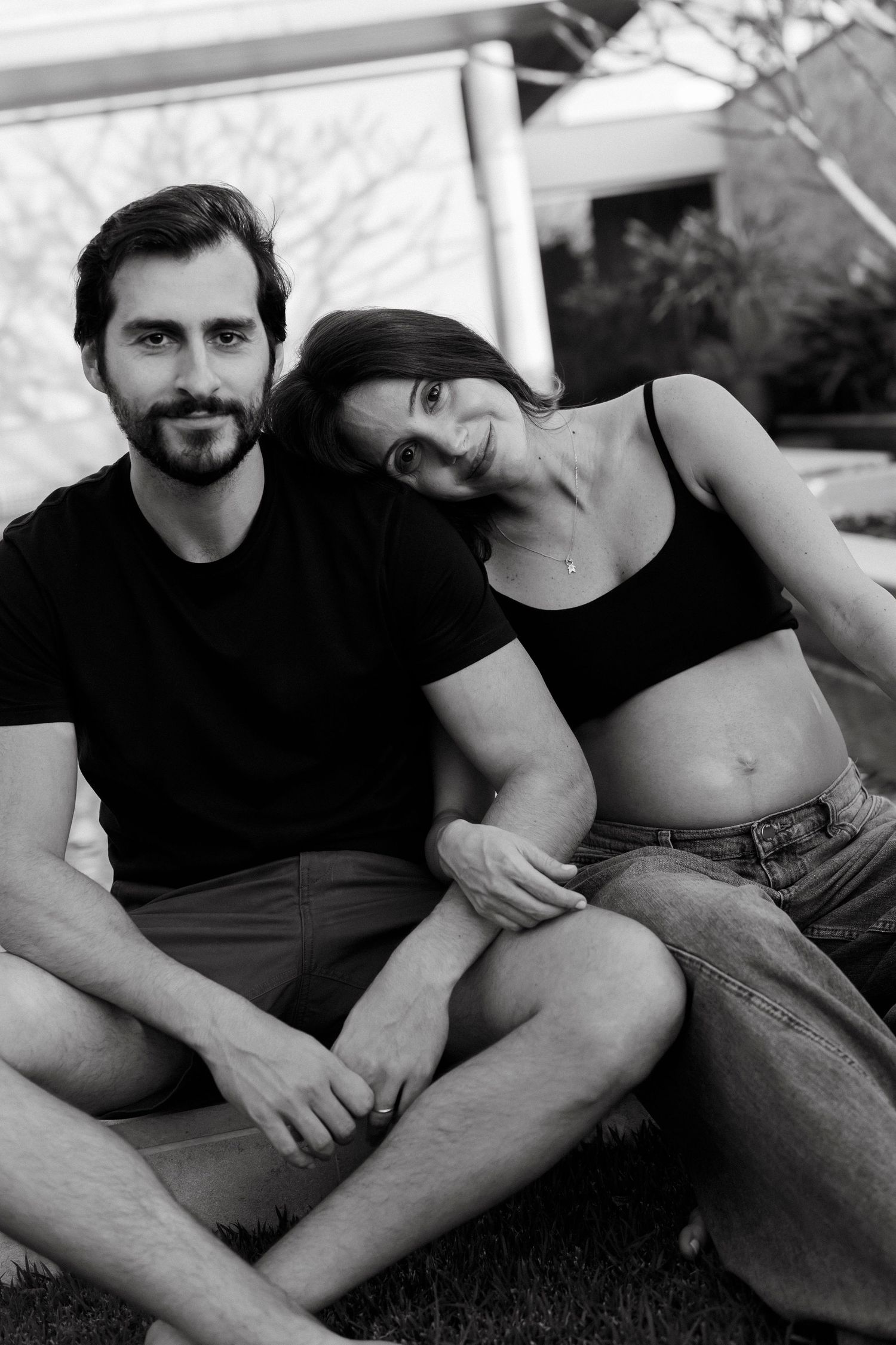 Black and white photo of a smiling couple sitting together on grass outdoors, woman leaning on mans shoulder.