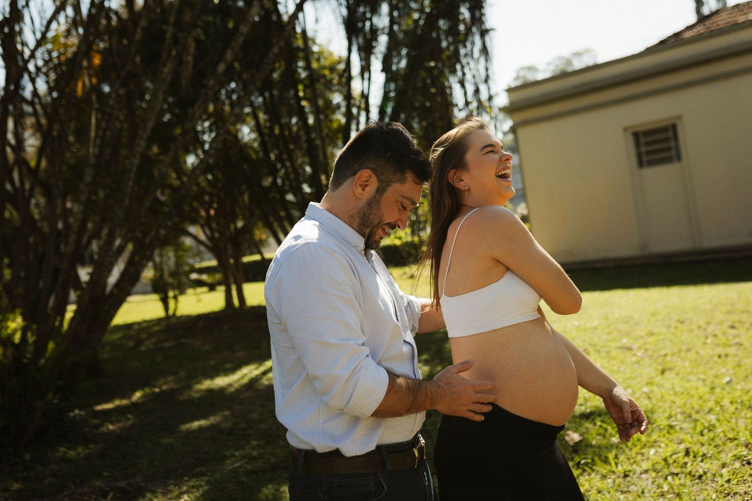 Pregnant couple laughing outdoors in sunlit garden, man in white shirt embracing woman showing baby bump.