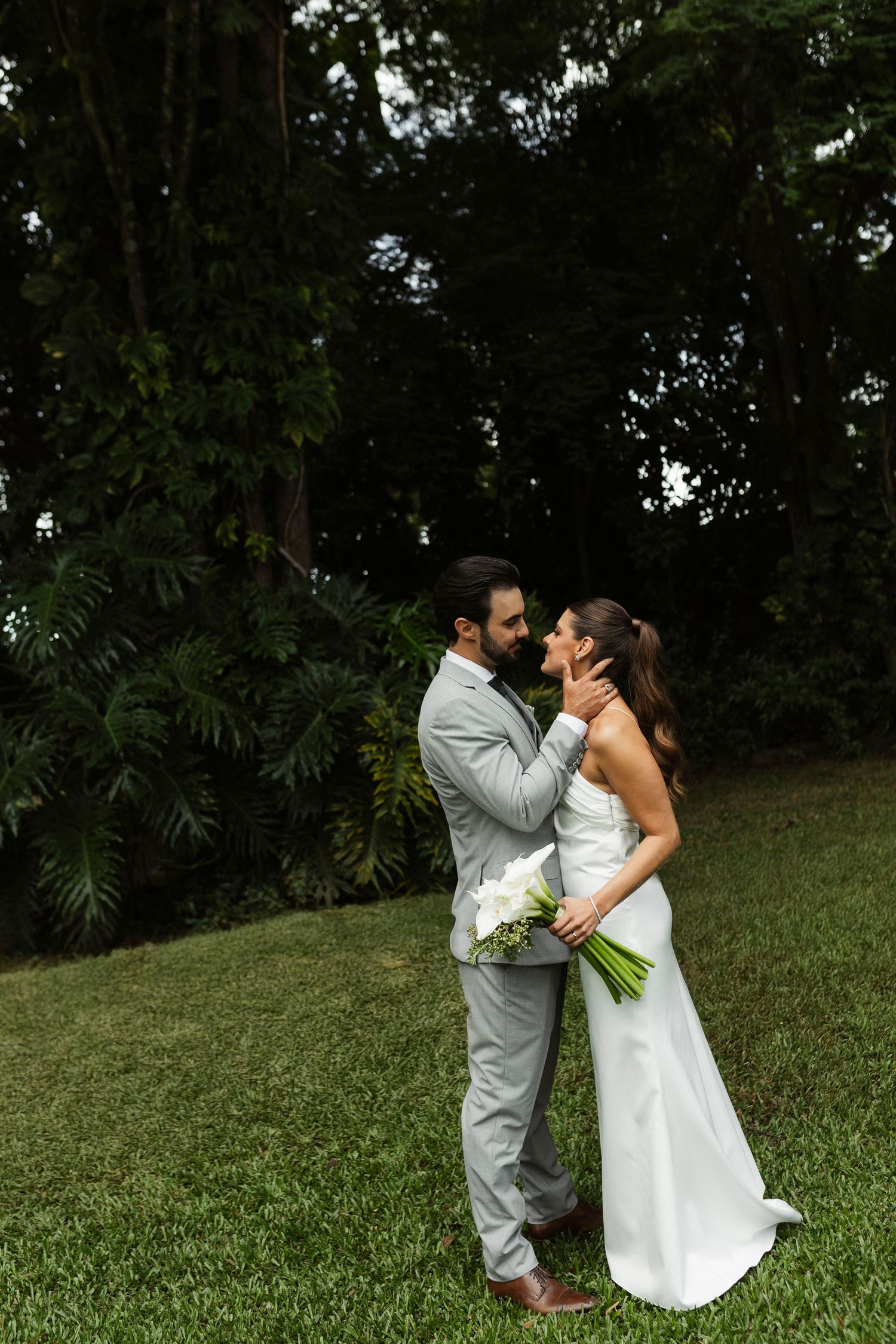 Bride and groom share a romantic kiss outdoors on green lawn with lush tropical trees in background.