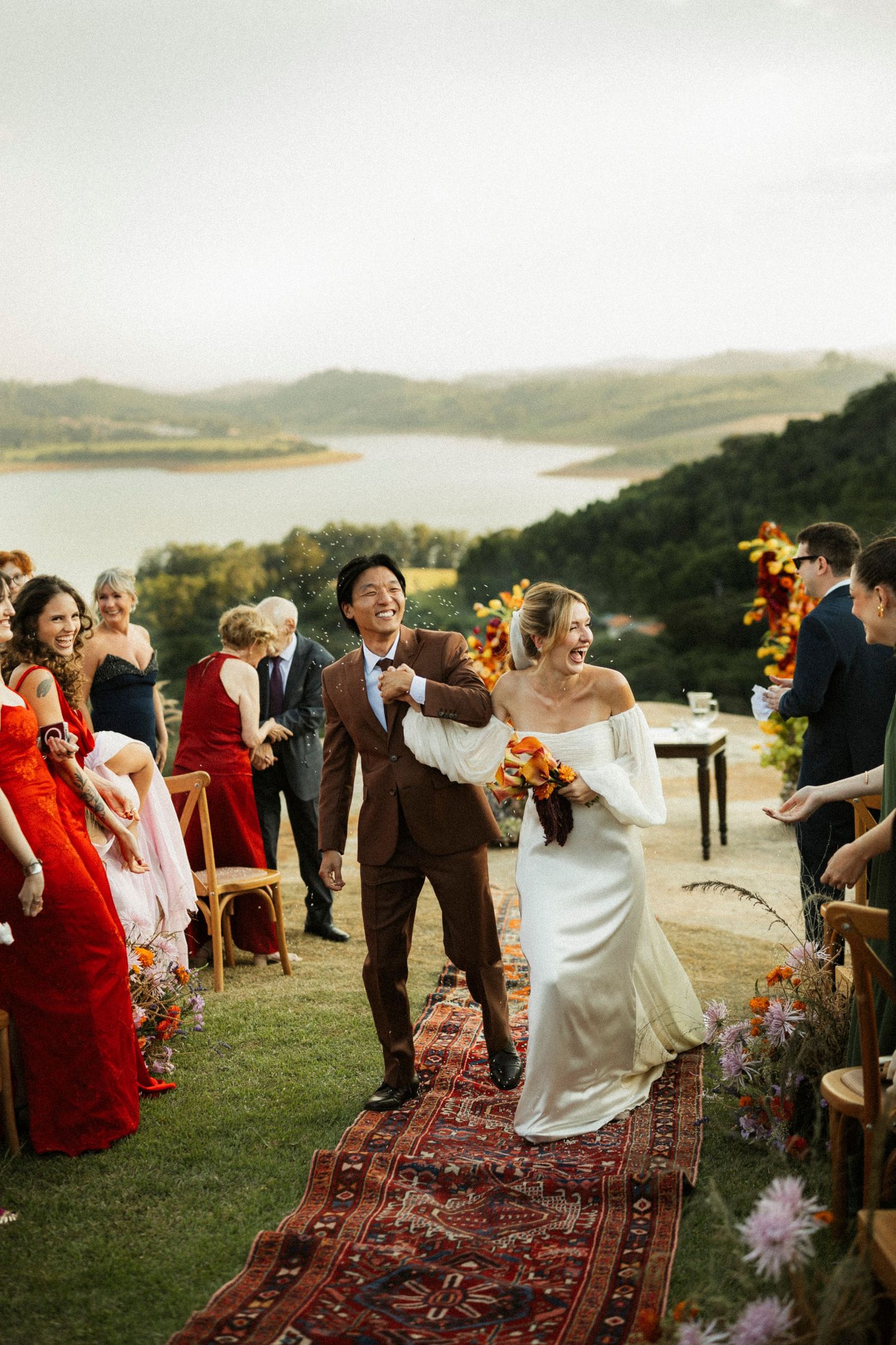 Joyful newlyweds walk down outdoor aisle on ornate rug with lake views and guests in colorful attire.