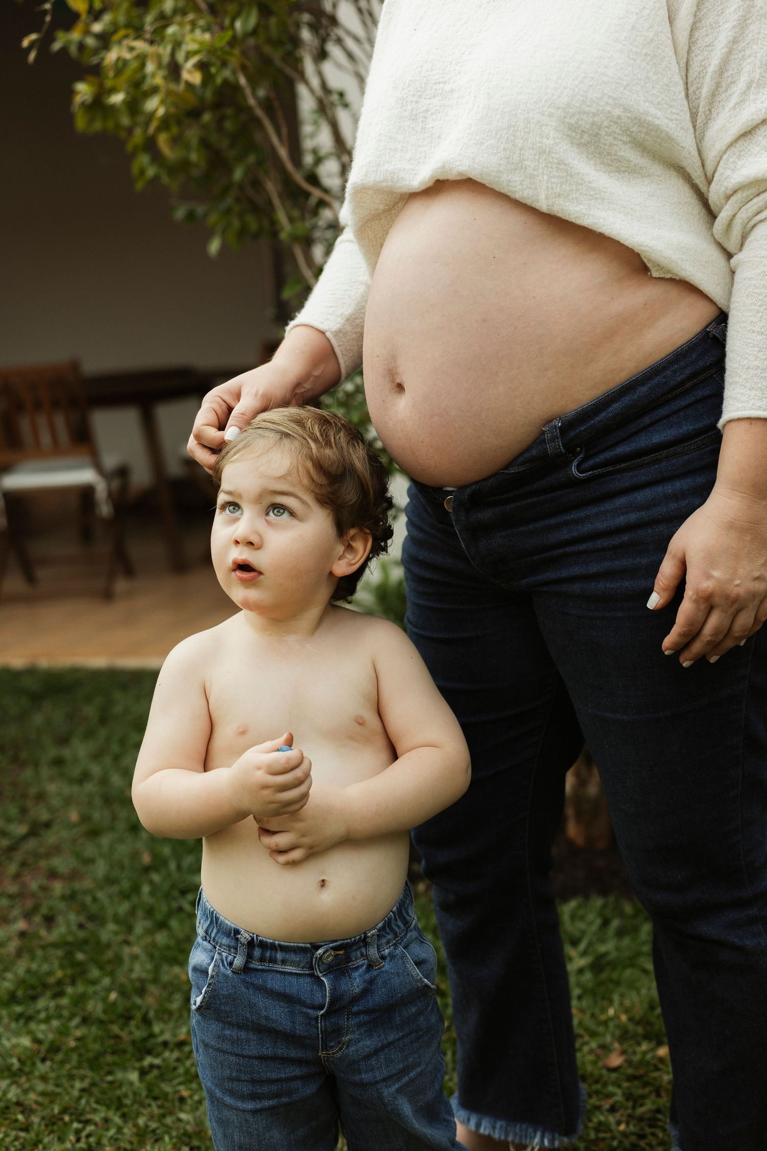 Toddler boy in jeans stands shirtless beside pregnant mother outdoors, looking up curiously as she touches his hair.