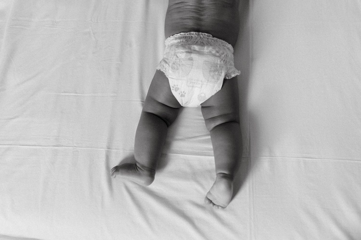 Black and white photo of a baby lying on a bed wearing a diaper, showing chubby legs and back.
