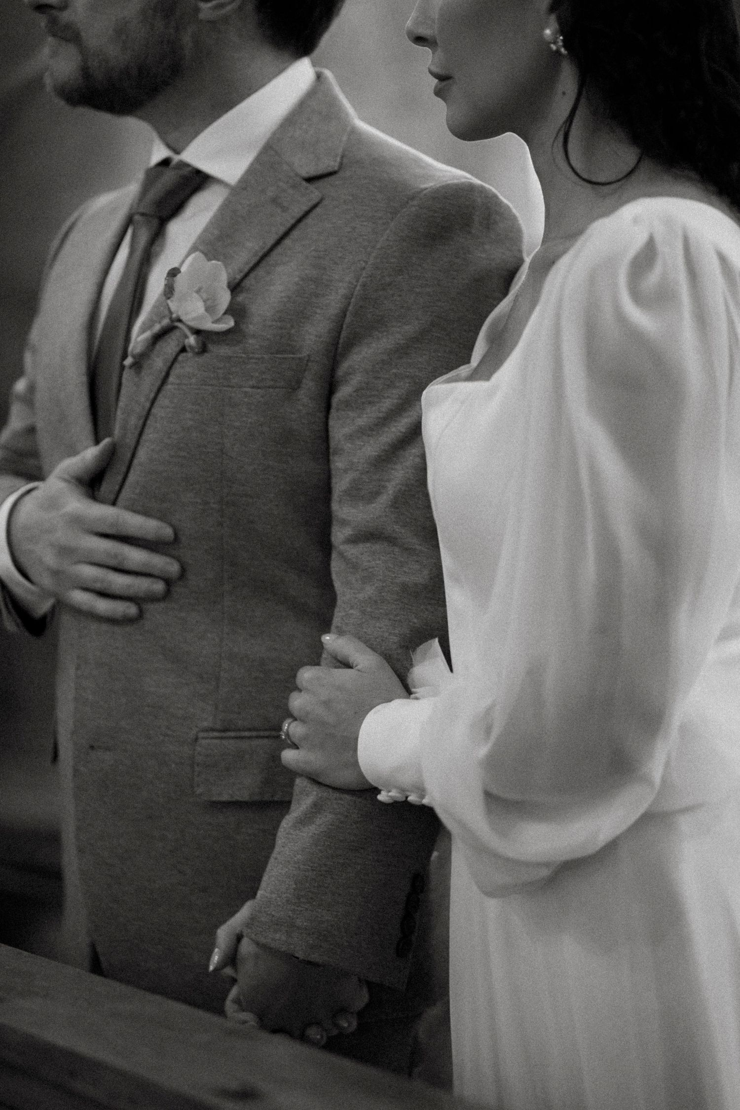 Black and white photo of a bride in white dress holding groom's arm in gray suit with boutonniere at wedding ceremony.