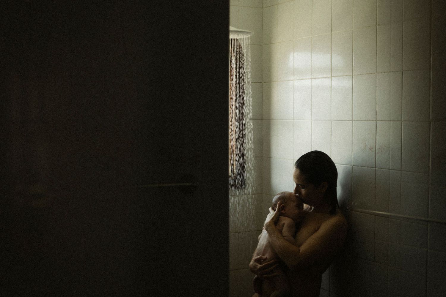 A mother holds her newborn baby in a dimly lit bathroom with white tiles.