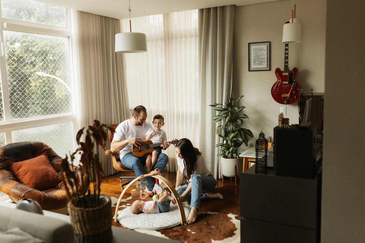Family enjoying music together in a cozy living room with guitar, plants, and warm decor.