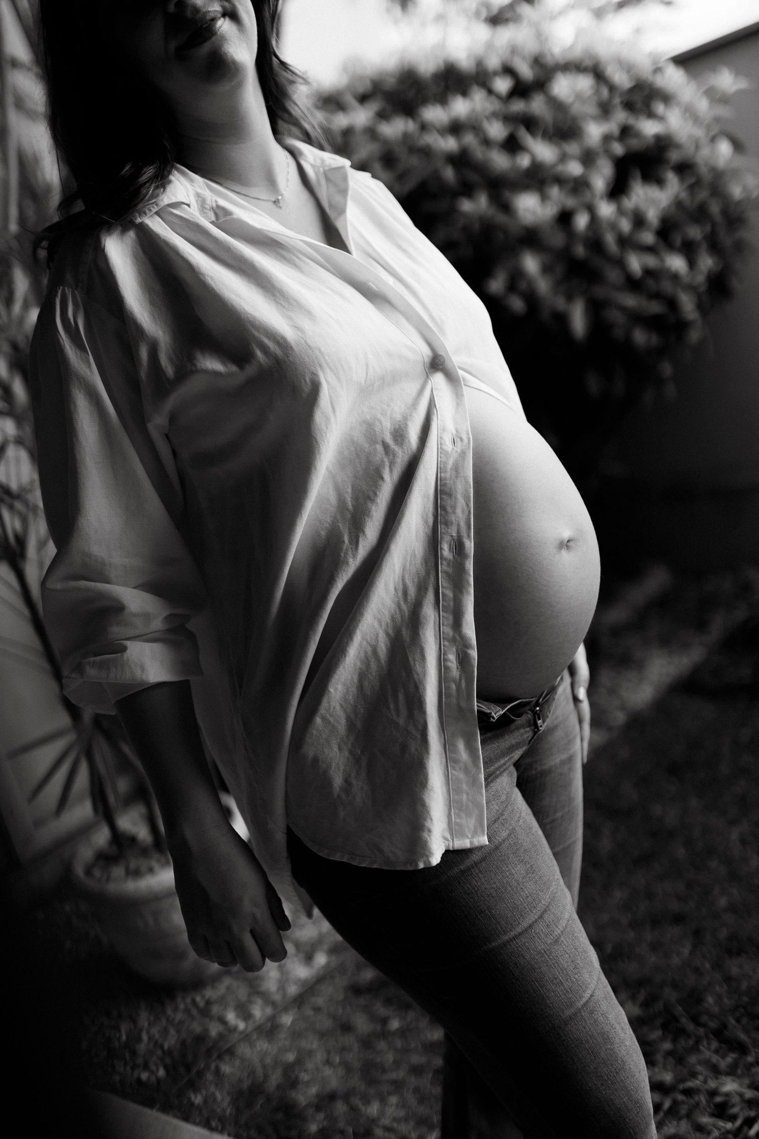 Black and white photo of a pregnant woman in an open white shirt and jeans, showing her baby bump outdoors.