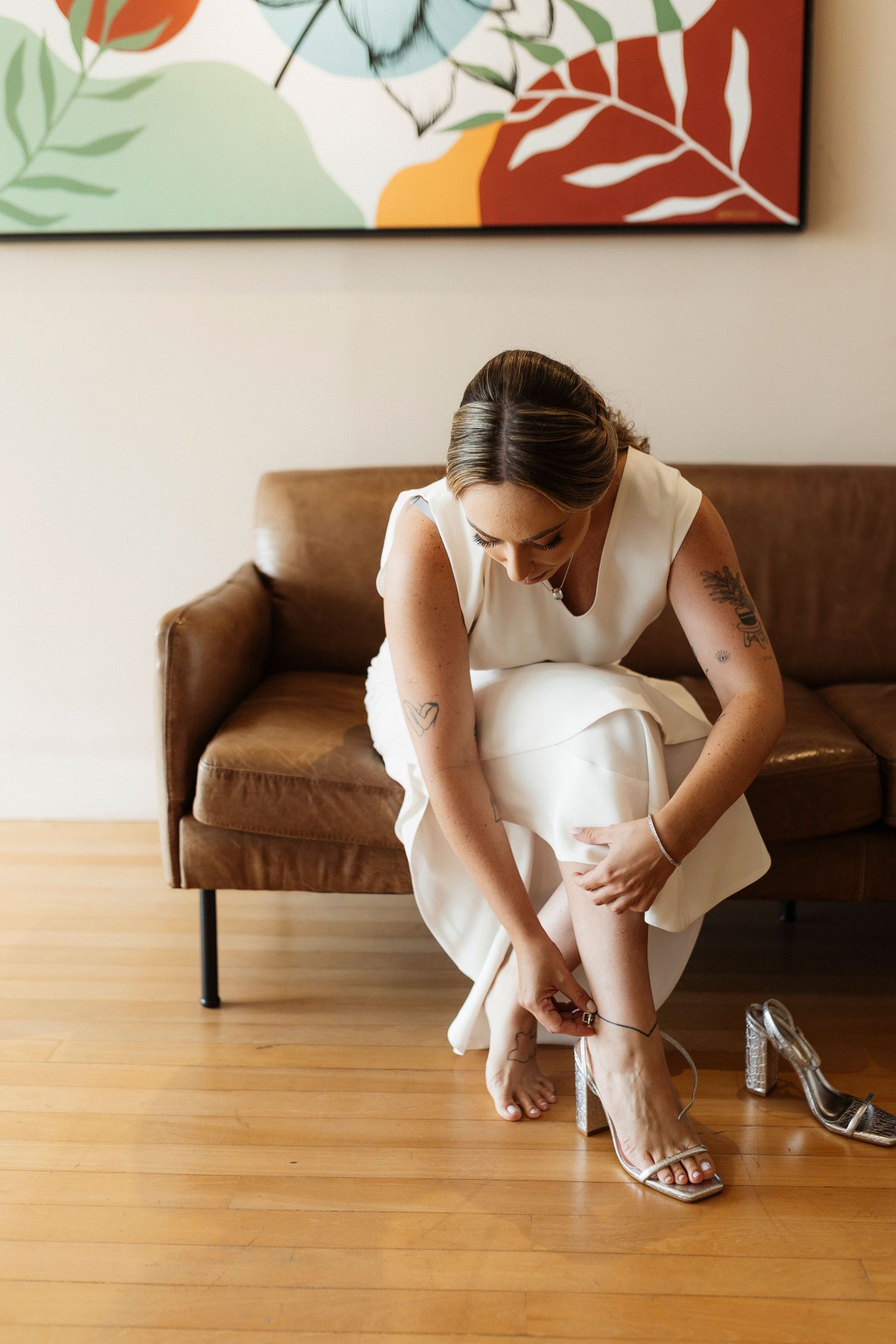 Bride in white dress adjusts her sandal while sitting on brown leather couch beneath colorful artwork.