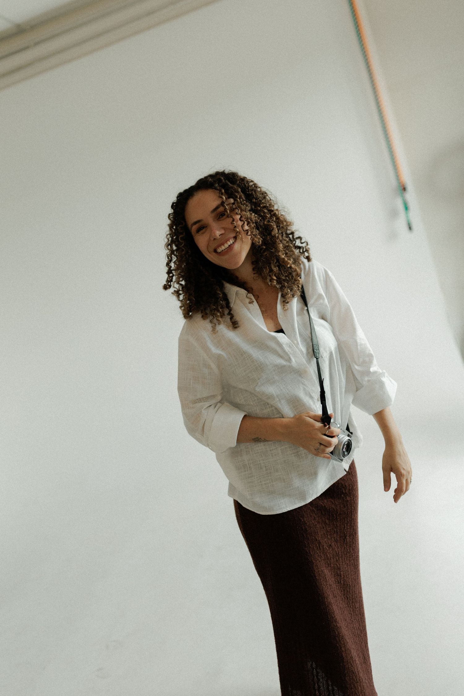 A figure in a white button-down shirt and brown skirt poses playfully against a white wall in a photo series.