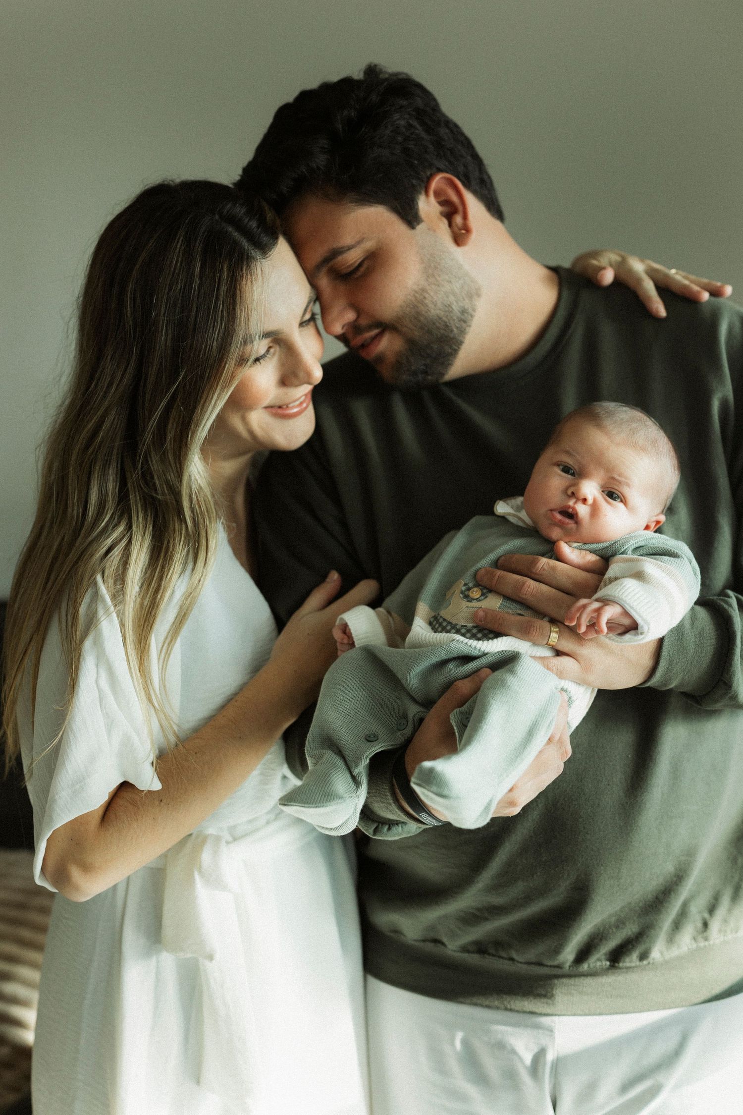 A young couple smiles lovingly at their newborn baby, with the mother in white and father in olive green.