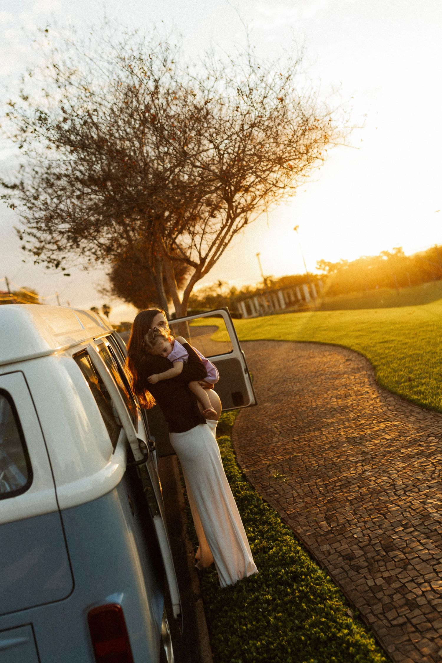Woman leaning out of a vintage van door at golden sunset, with a winding brick path and tree in the background.