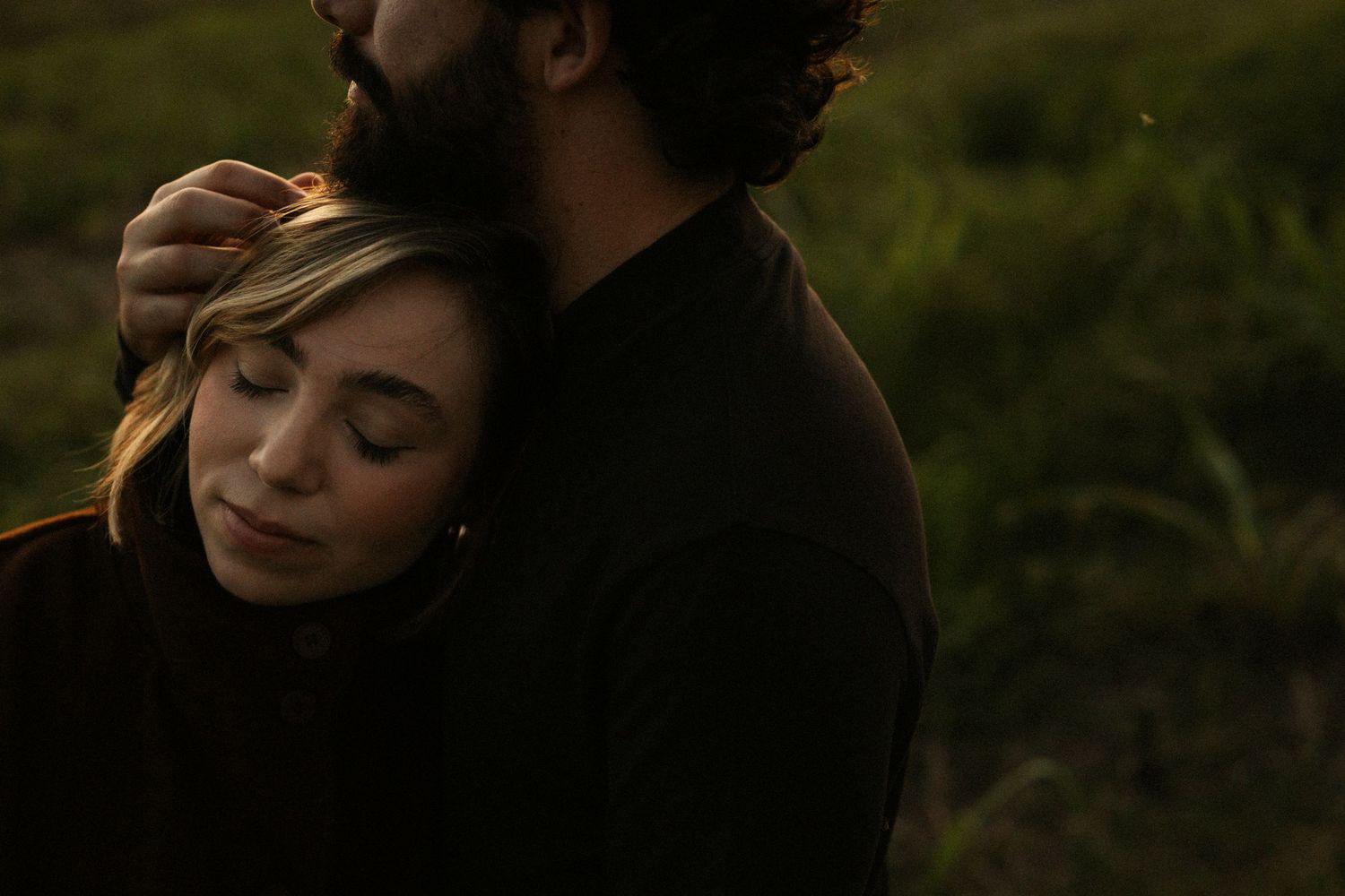 A woman with blonde hair rests her head on a bearded man's chest outdoors in a tender romantic embrace.