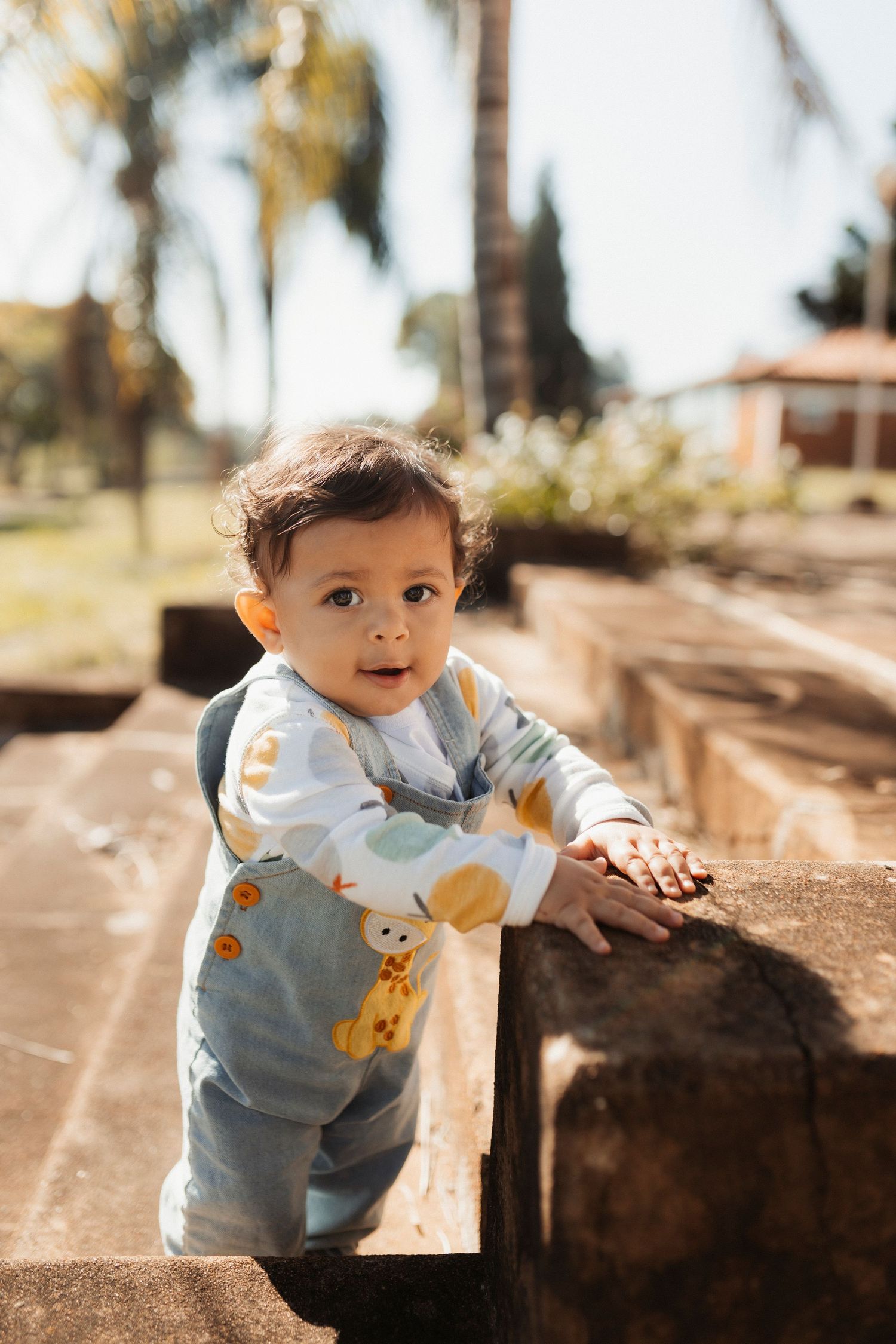 Baby in light blue outfit exploring outdoor setting with palm trees.