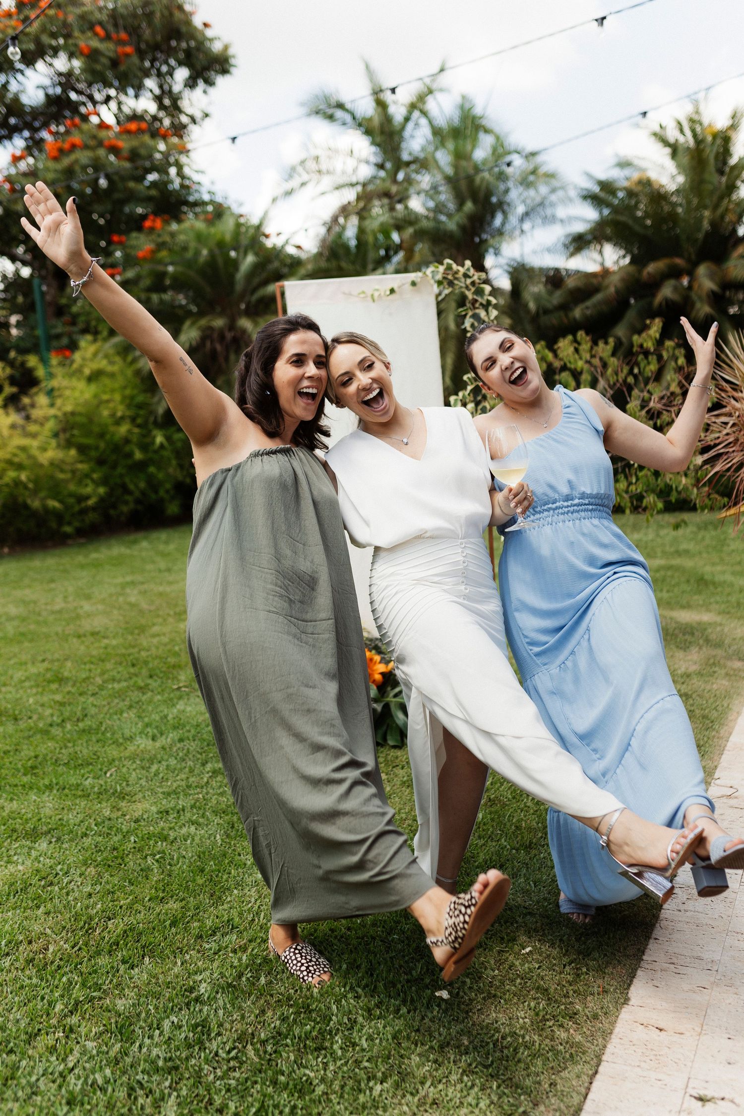Three friends in casual clothes laughing and jumping together on a green lawn.