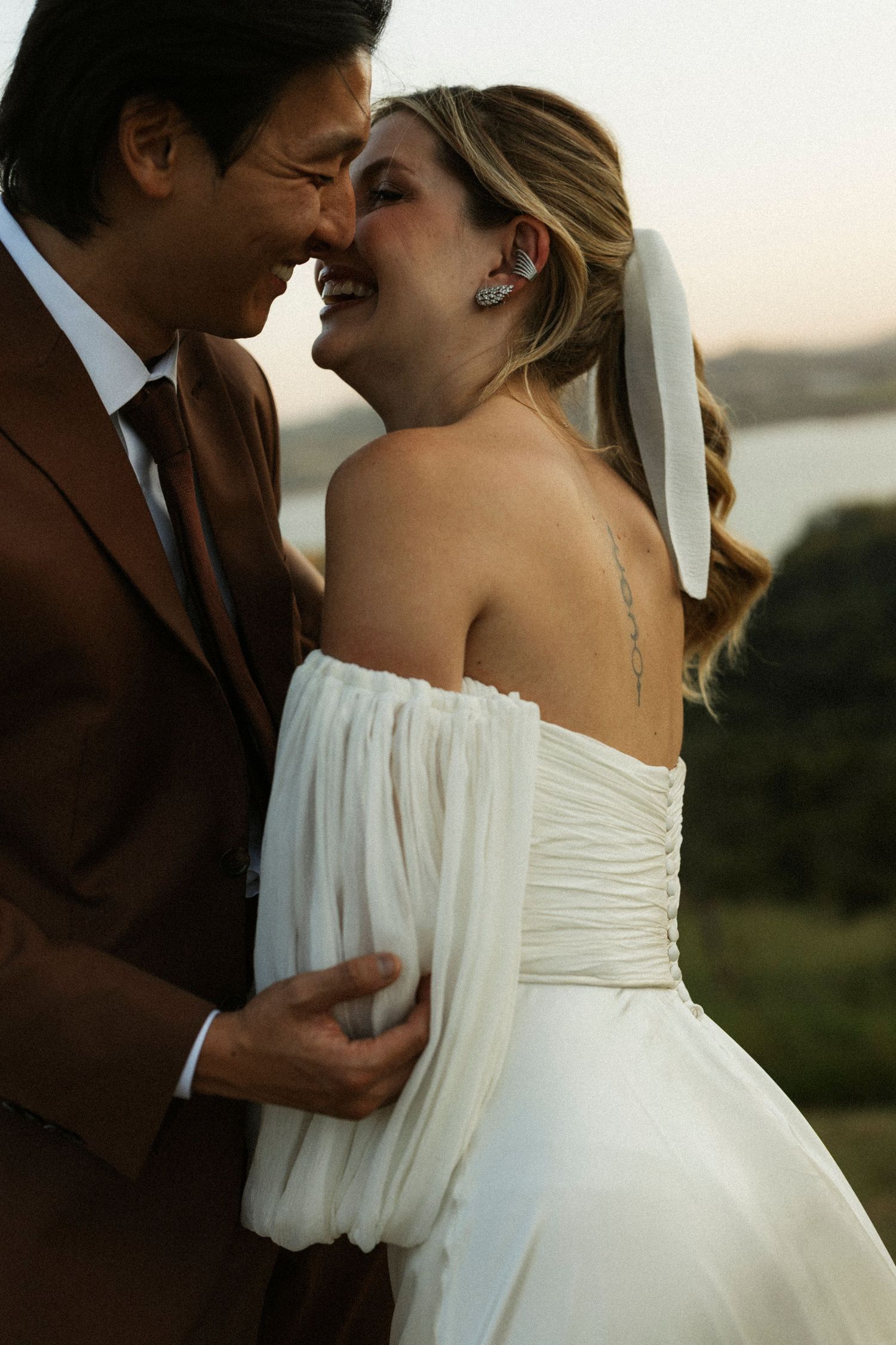 A joyful bride in a white off-shoulder gown and groom in a brown suit share a tender moment outdoors.