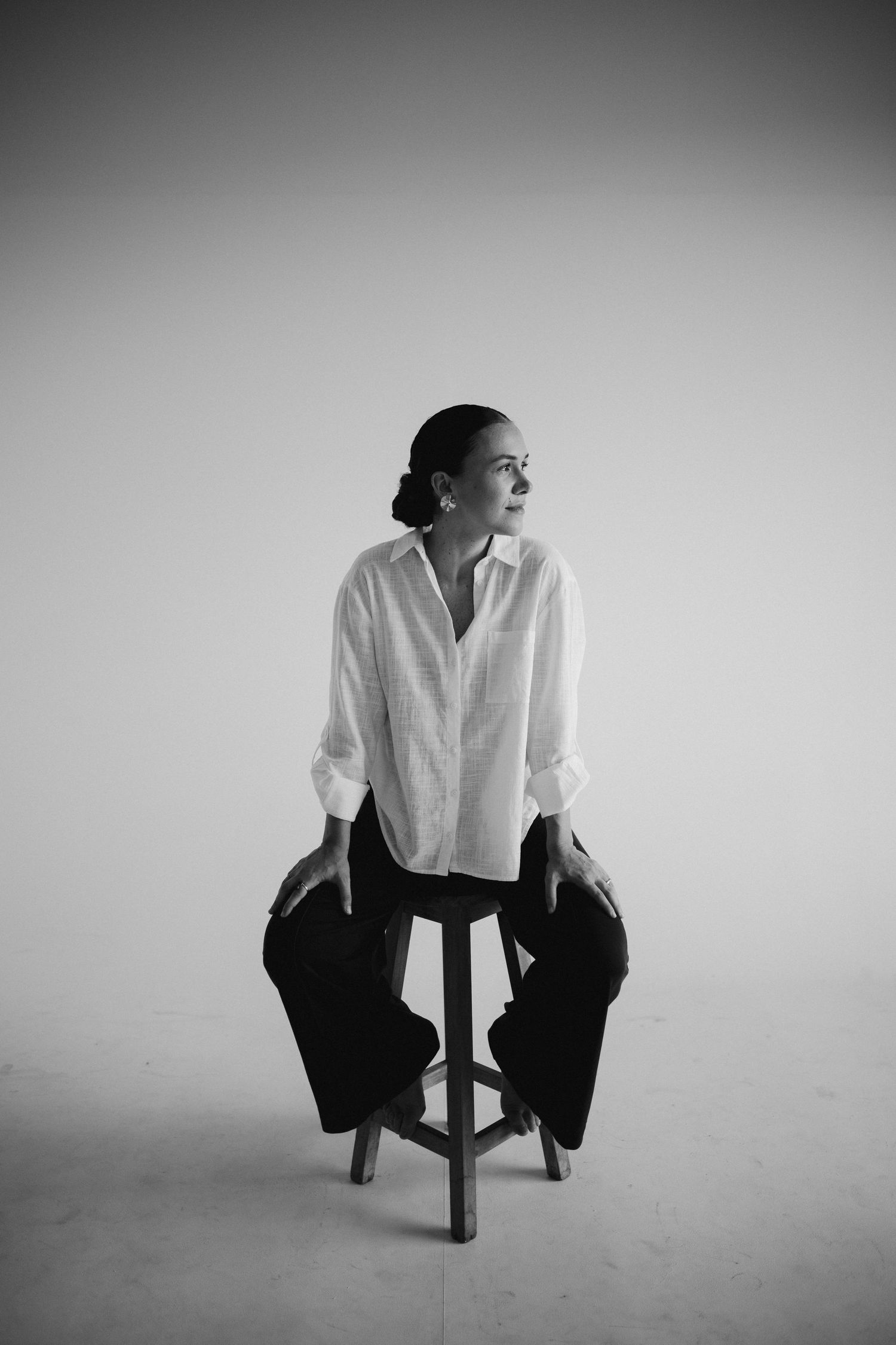 Black and white portrait of someone sitting on a stool wearing a white button-down shirt and dark pants in a minimalist setting.